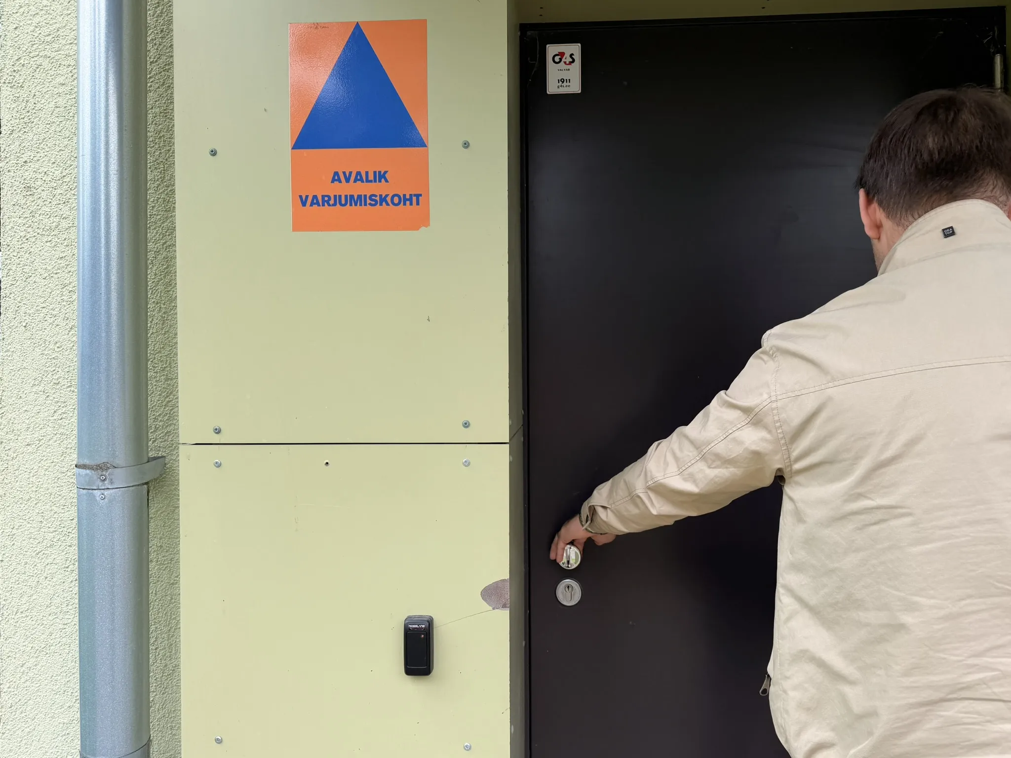  Mees beezes mantlis avab musta ukse heledal seinal. Oranžil plakatil on sinine kolmnurk ja tekst "AVALIK VARJUMISKOH", mis näitab avalikku varjupaika.