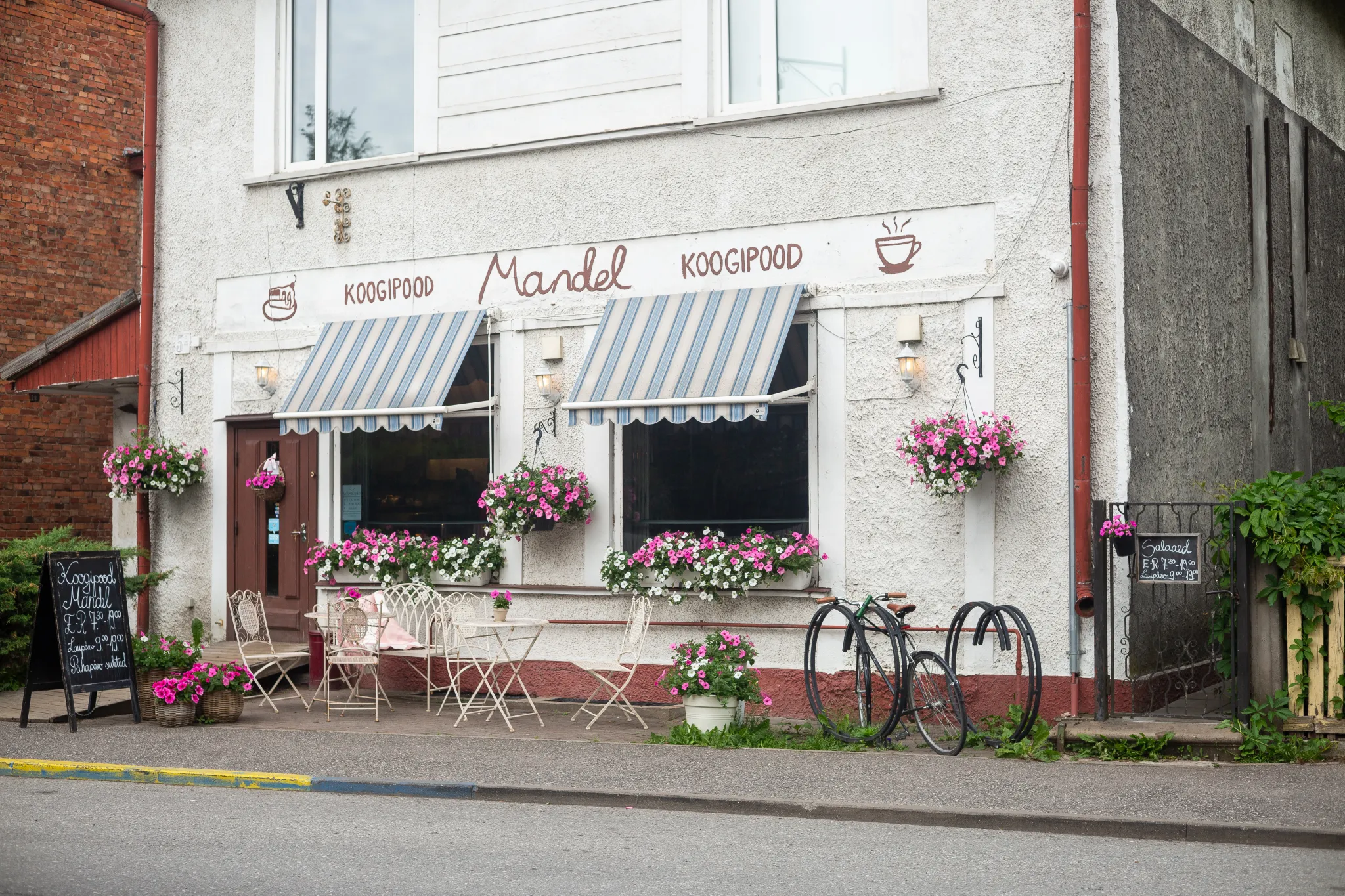  Väike pagarikohviku ettevaade: sinivalge triibulised rulood, valged välilaudade ja toolide komplekt, rippuvad lillekorvid. Kaks jalgratast ukse ees; tänavaposti tahvel menüü. Silt: Mandel Koogipood.