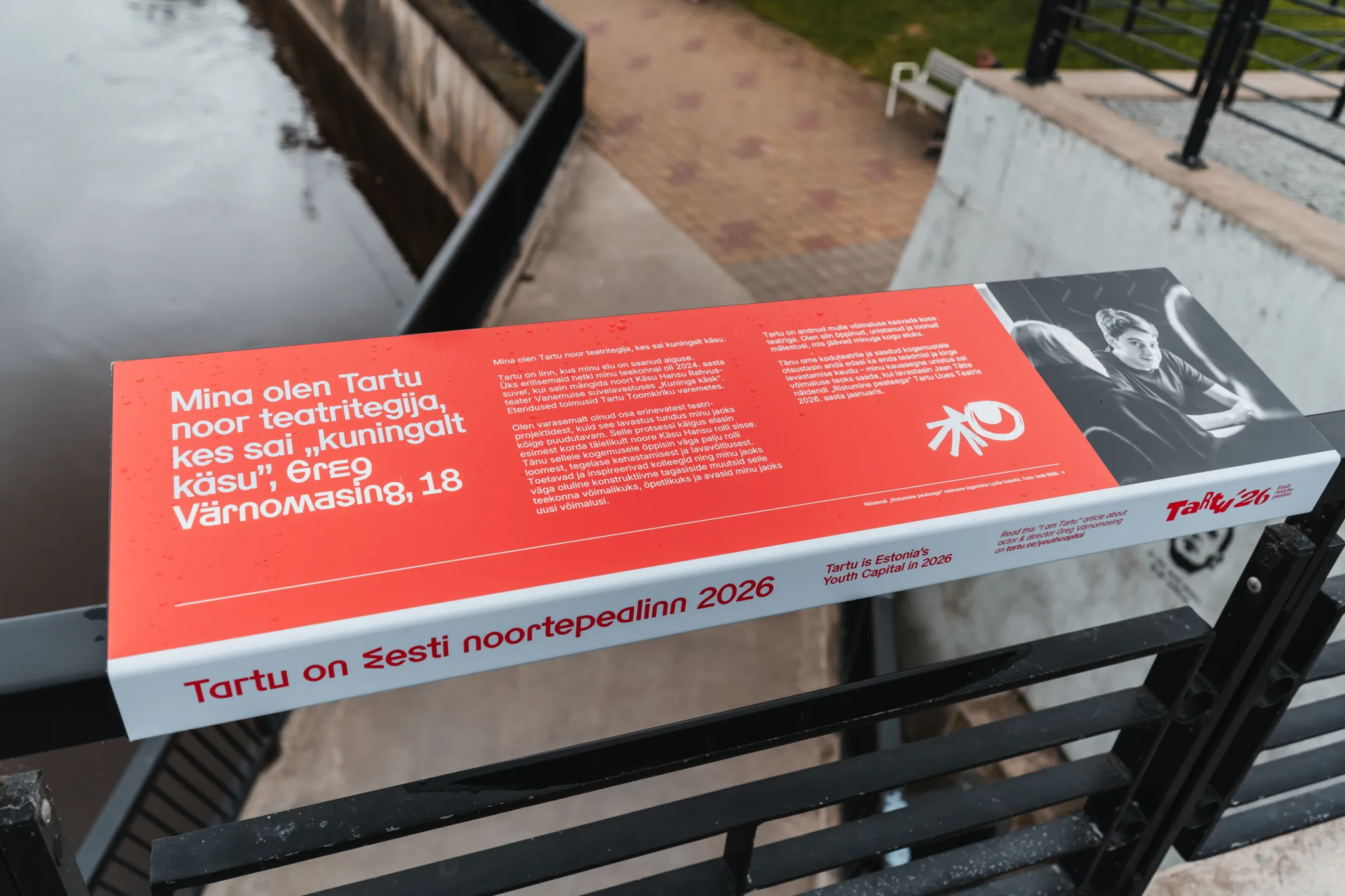  Punane banner käsipuul Tartus, kanali ääres, reklaamib Tartut Eesti noortepealinn 2026; sisaldab teksti ja mustvalget fotot.