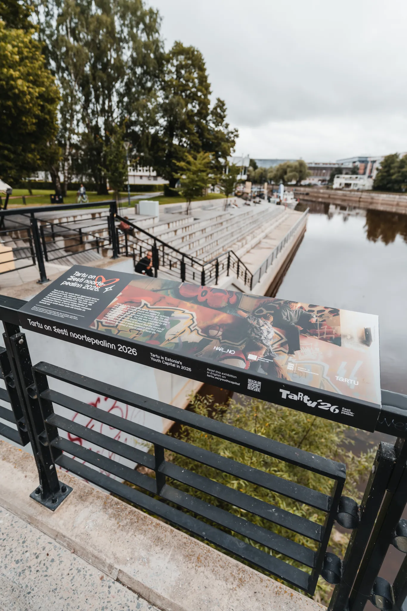  Tartu jõekallas asuv promenaad musta metallraamiga aiaga, trepid vete äärde, rohelised puud ja kaugel linn. Aia ääres pikk plakat reklaamib kohaliku üritust.