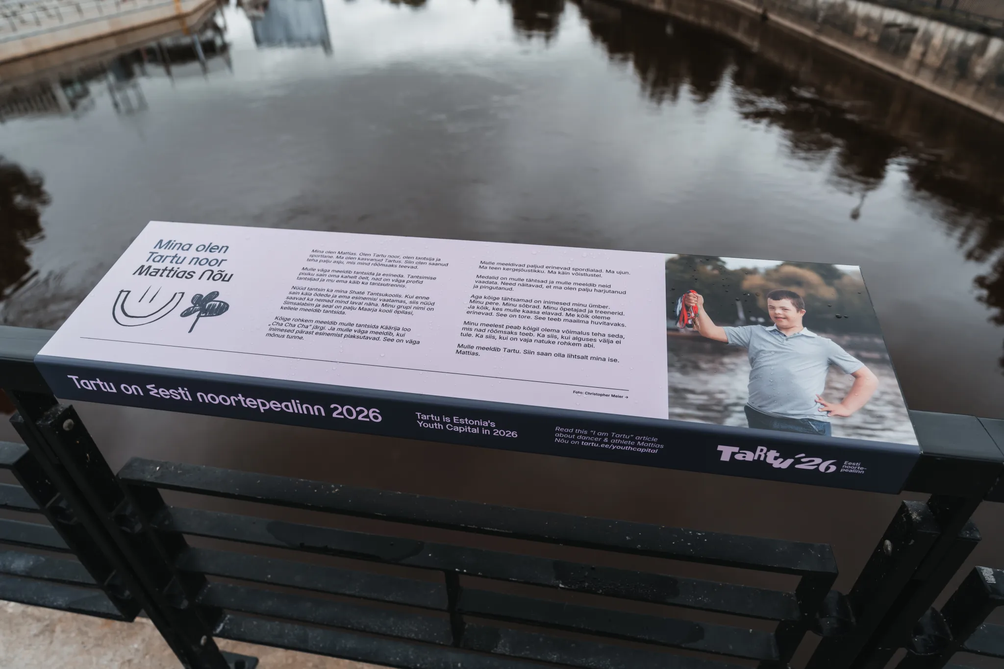  Infoplakat Tartu kanali ääres, Eesti; valgel paneelil poisi foto ja tekst Noortepealinn 2026 kohta, taustal rahulik kanal ja peegeldused.
