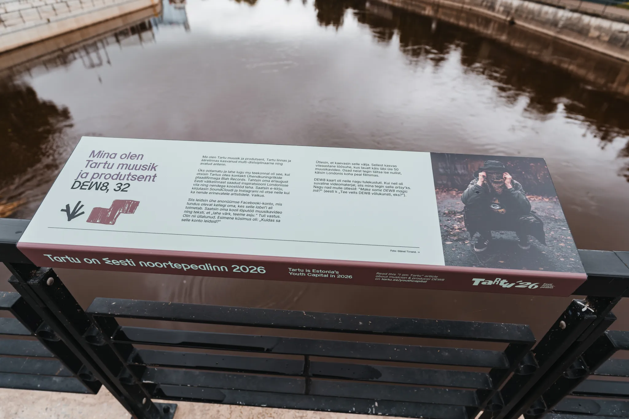  Pikk infoplaat istme kõrval jõe kaldal Tartus, Eestis. Paneelil on tekst ja foto ning teave Tartust noortepealinnana 2026. Taustal on vesi, kai ja kajastuv pind.