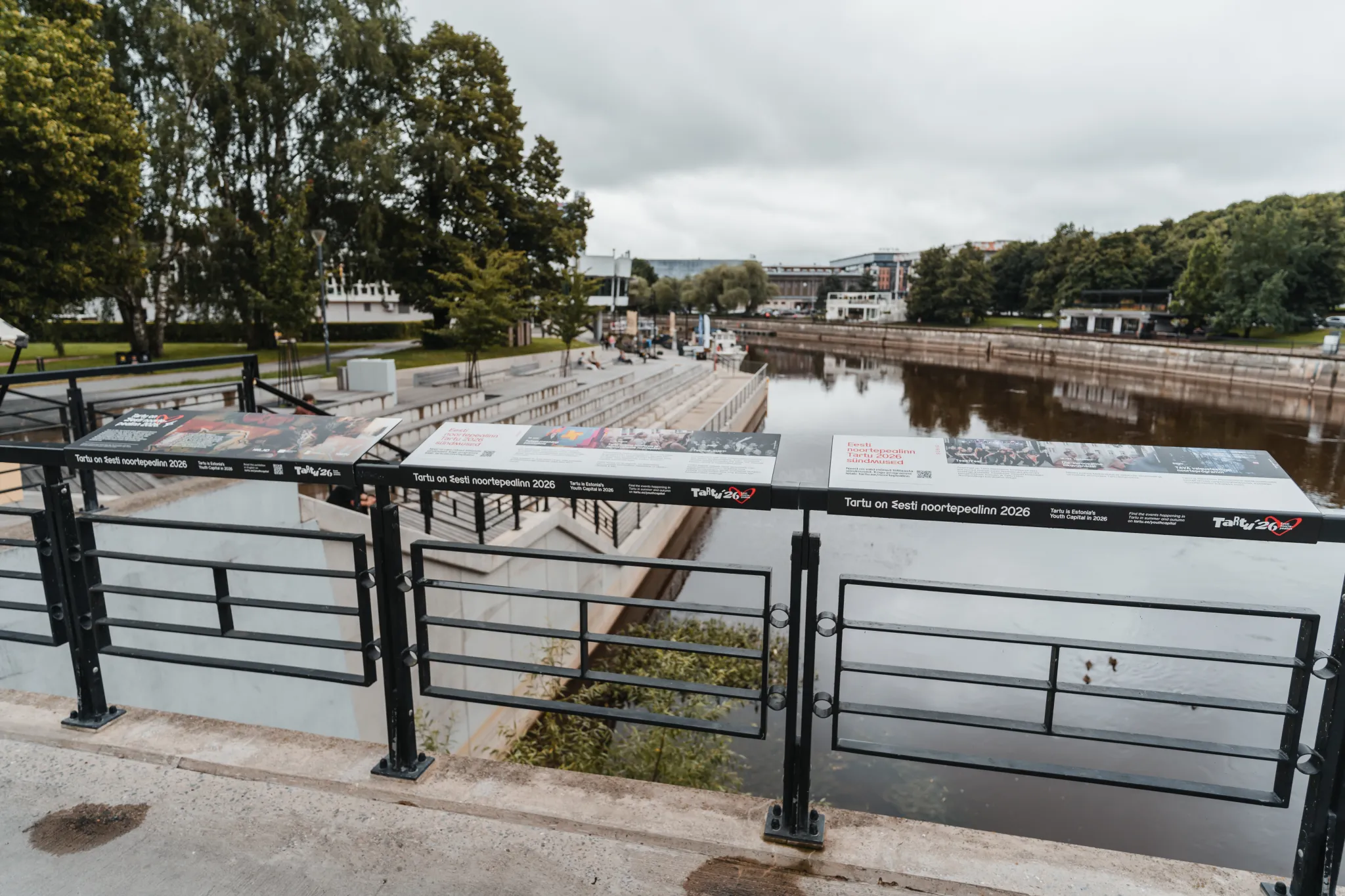  Tartu jõeäärne promenaad Emajõe kaldal, kaasaegsete mustade piirdega, kivised sammud vee äärde, puude ja pargiga ning inimestega jõe ääres; pilves taevas ja kaugel linnamajad.
