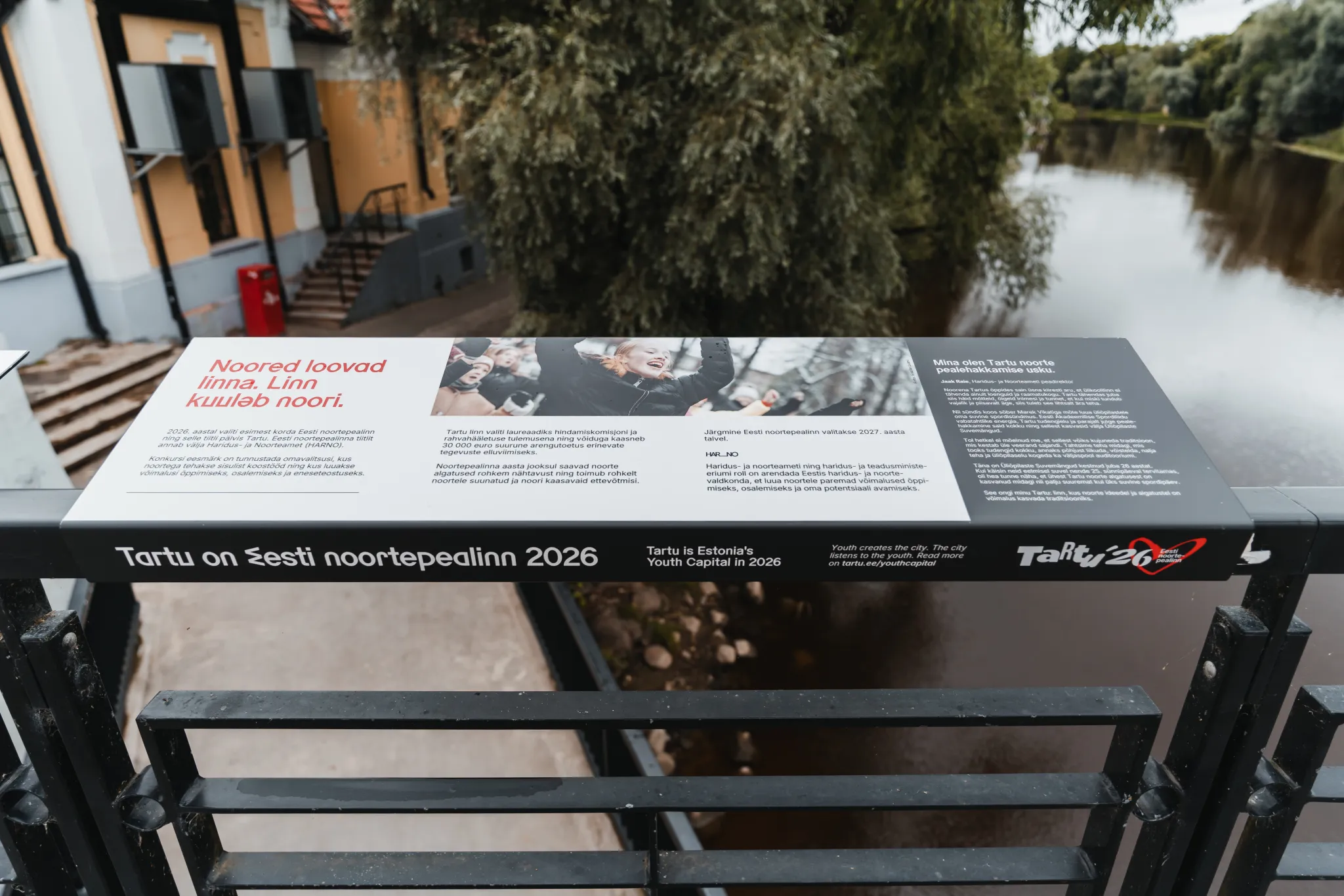  Infostend silla raami kohal kanali ääres Tartus, Eestis. Plaat kuulutab: "Tartu on Eesti noortepealinn 2026" koos fotode ja tekstiga, lähedal oranž hoone, trepid ja rahulik veekallas puude taustal.