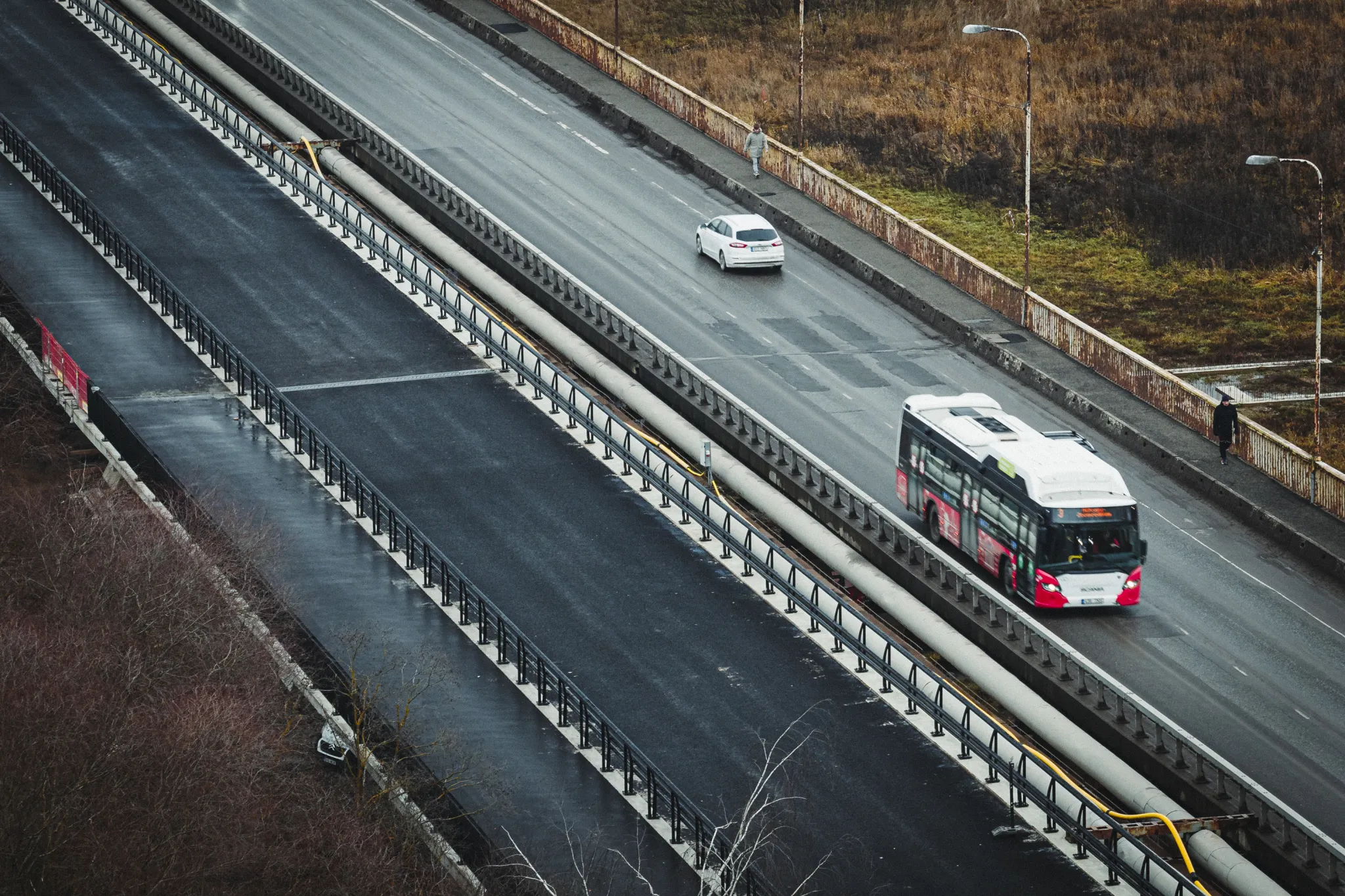  Õhust vaade moodsale linnakiirteele; keskbarjäär eraldab sõiduteed. Ühes reas sõidab punase-valge linnabuss, teises reas valge auto; teeääri piirab aiaga.
