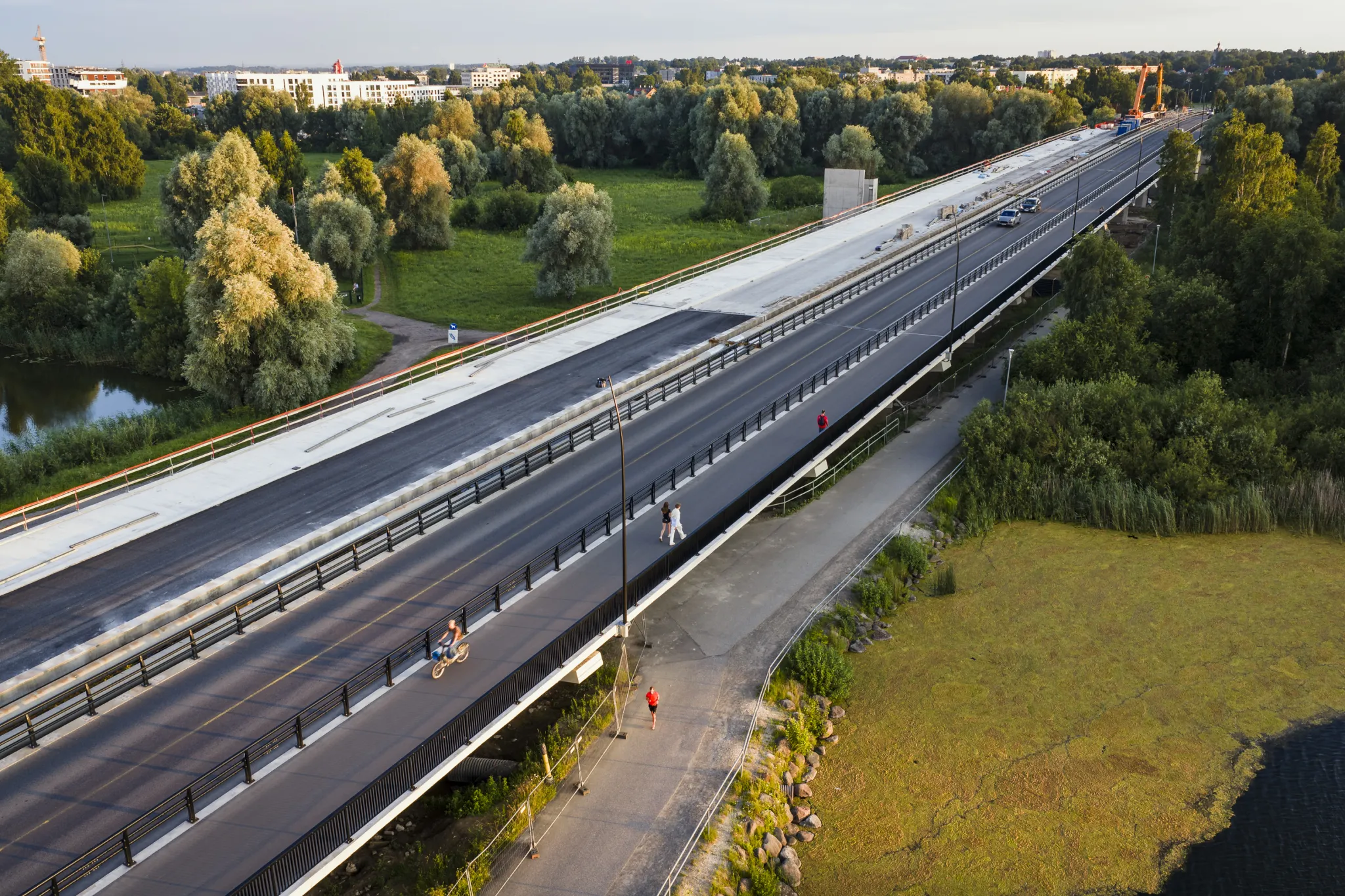  Õhust vaade pikkale sillale Tartu lähedal, kõrvalt laiad pargid ja veeteed. Keskosas sõidurajad autodele, ääres jalakäija- ja jalgrattateed. Inimesed sõidavad ja kõnnivad; kalda ääres puud ja vesi.