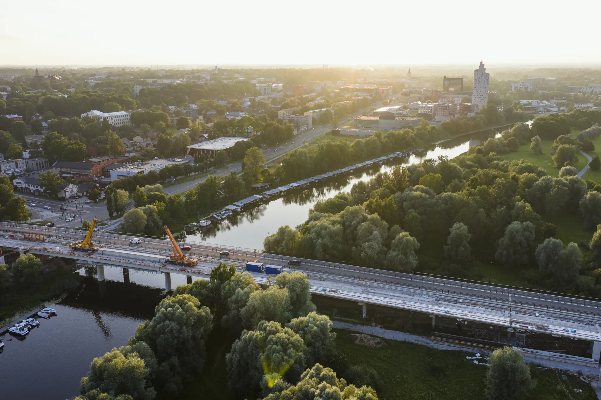  Õhust vaade Tartu linnale: jõgi, rohelised pargid ning ehituskraanidega sild päikeseloojangul.