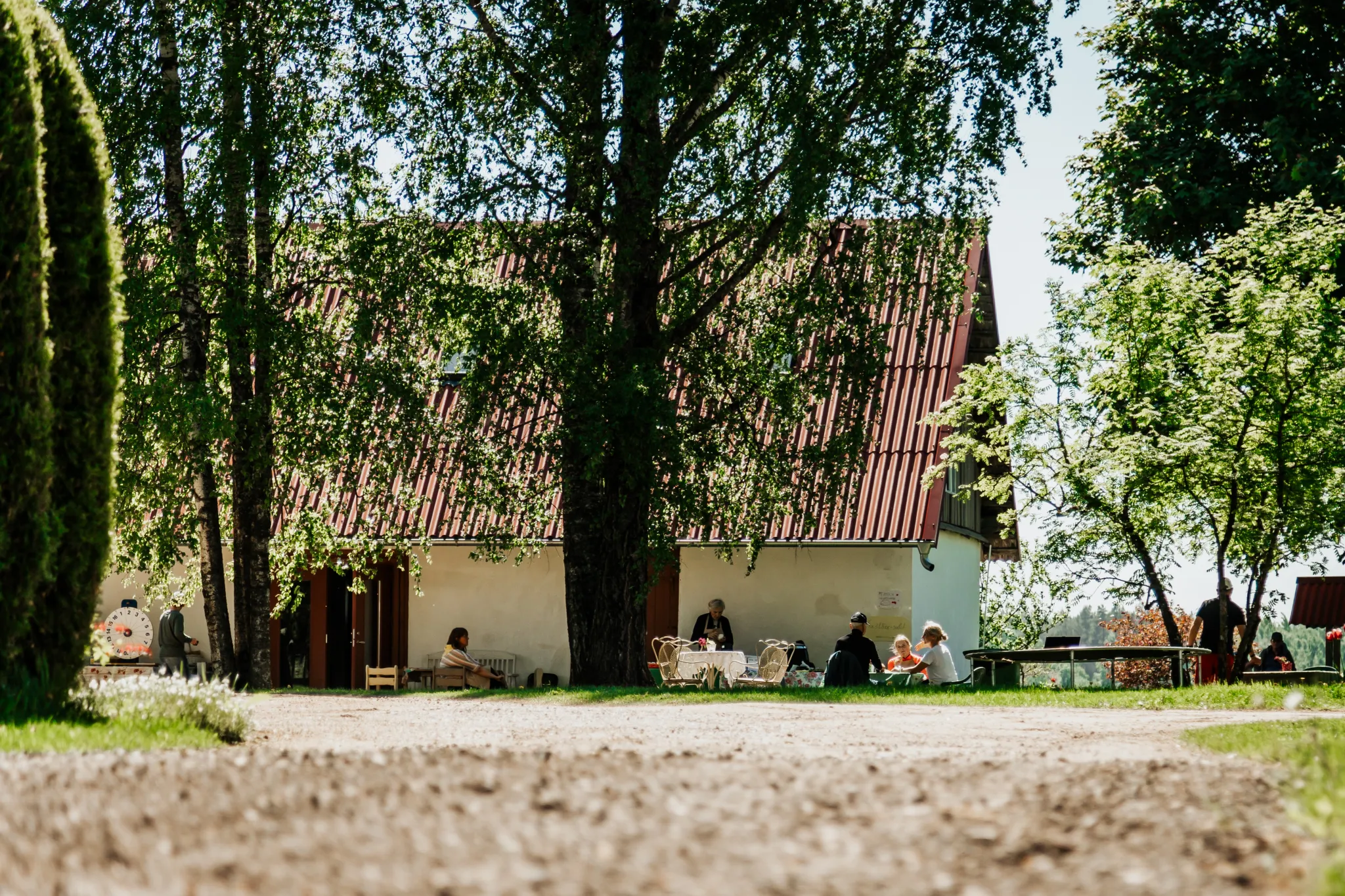  Varjus pargis on valge hoone punase katusega. Puude all istuvad külastajad ja naudivad väliterrassi.