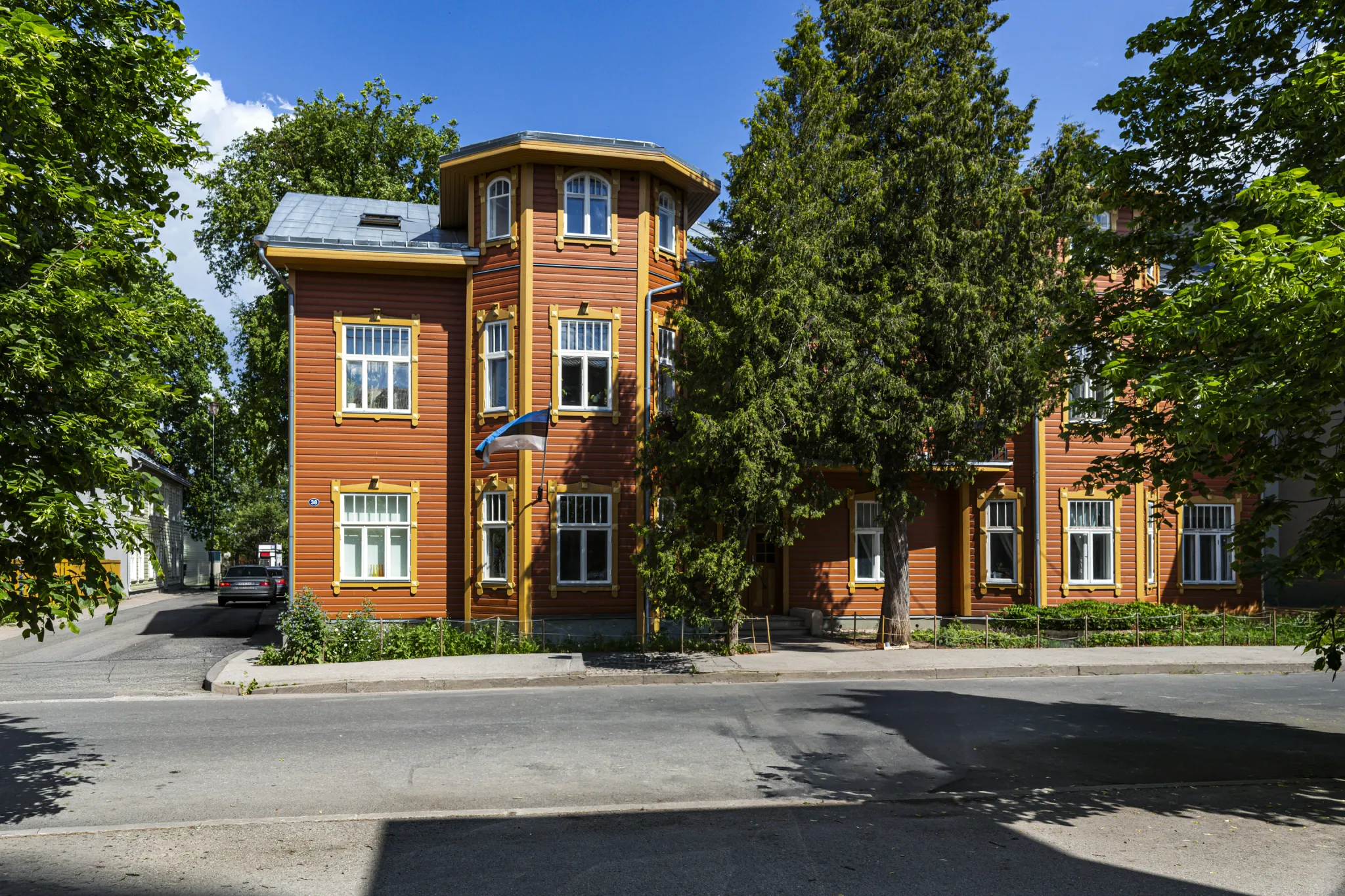 Oranž puidust maja ümmarguse torniga, valged aknad ja sinine lipp. Suured puud ümbritsevad vaikset, päikeselist tänavat.