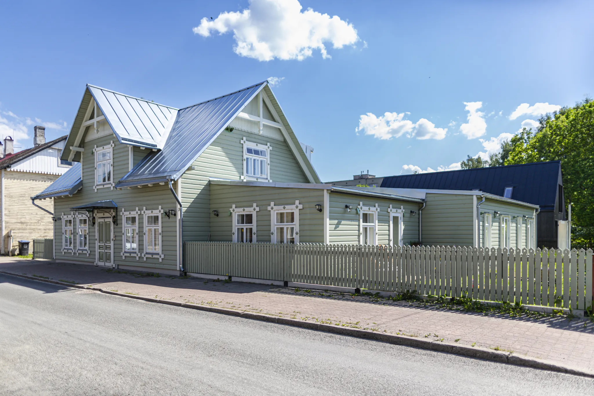  Roheline puidust maja valgete ornamentsete aknaraamidega, kelpkatusega ja väikese valge aiaga kõnnitee kõrval. Päikeseline tänav Eestis.