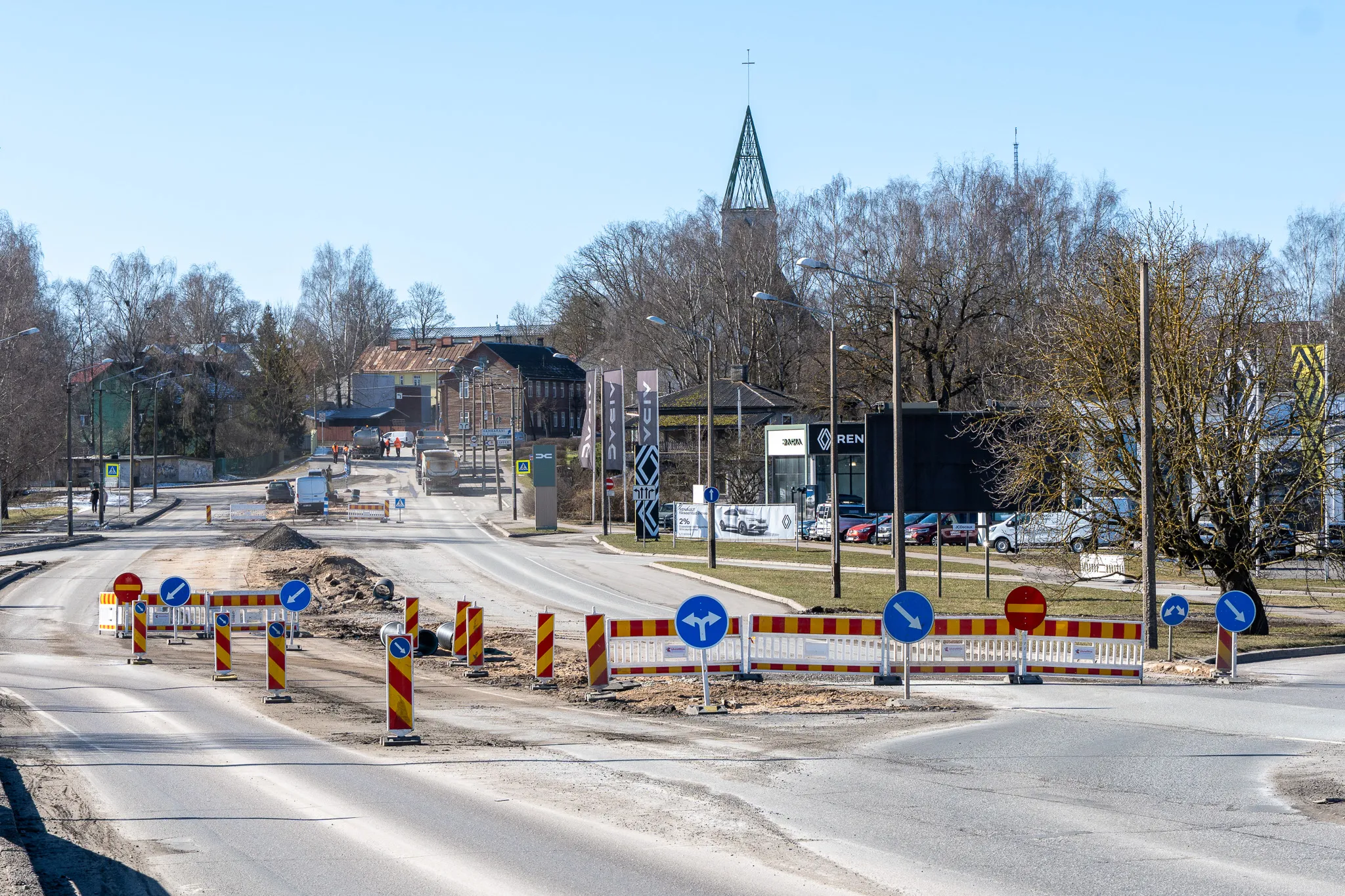 ** Vaade Tartu, Eestis, hõivatud teele, kus on mitmeid sõidukeid ja jalakäijaid. Teeäärtes on tänavatuled ning külgedel on nähtavad ehitustööd, mis viitavad käimasolevale arengule. Taevas on selge, mis viitab päikeselisele päevale.