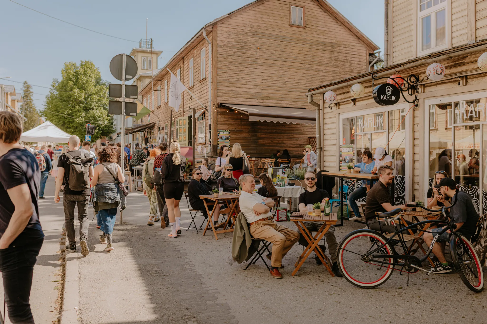  Päikeseline tänavakohviku vaade puidust hoonete vahel ja väliterrassiga. Inimesed istuvad ja vestlevad, jalgrattad pargitud lähedal, jalakäijad kõnnivad. Elav terrass ajaloolises Eesti linnas.