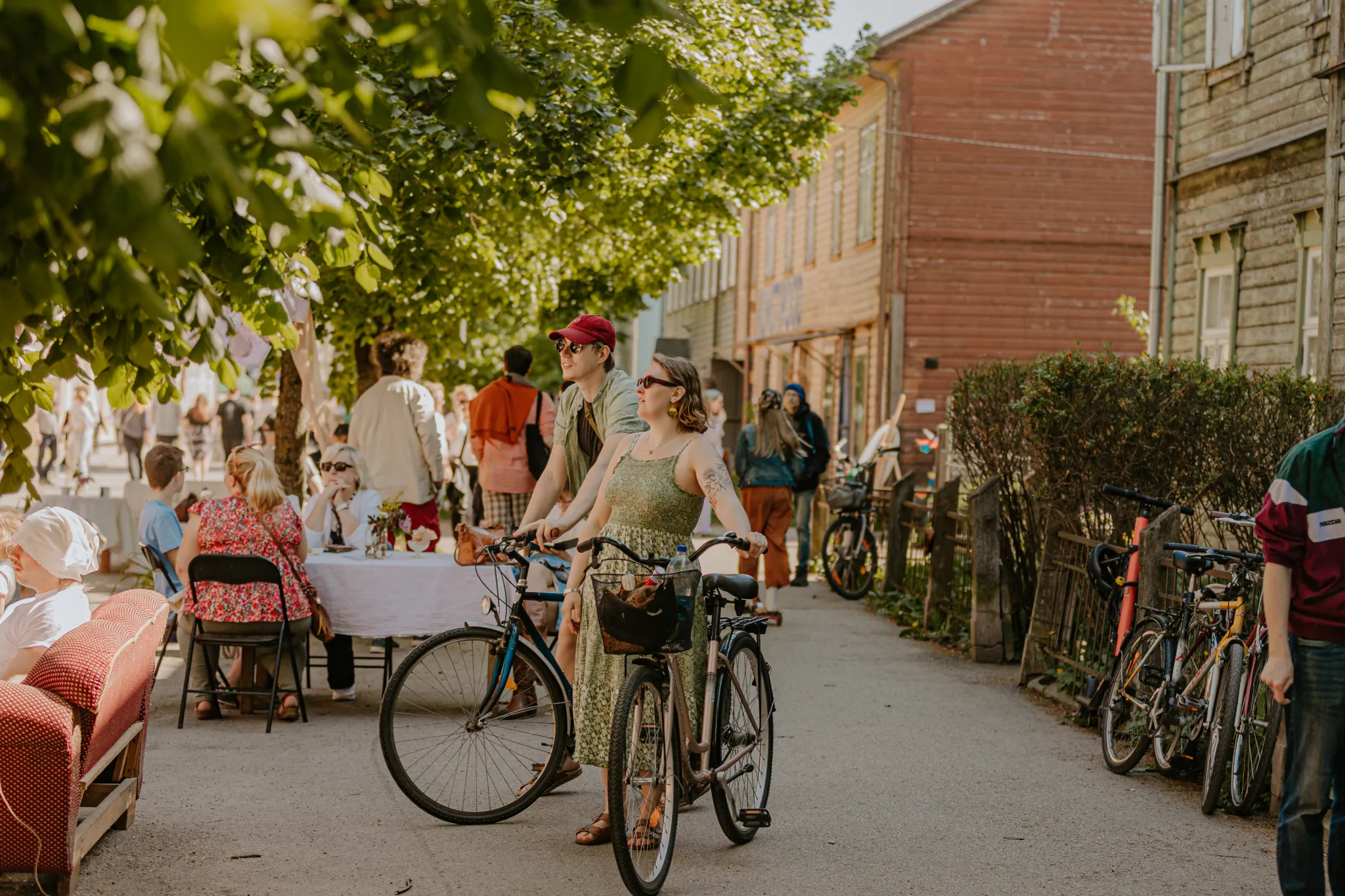  Päikeseline Tartu tänav: inimesed söövad välilaudade ääres, jalgratturid peatuvad ratastega ning varjul on ajaloolised puidust majad.