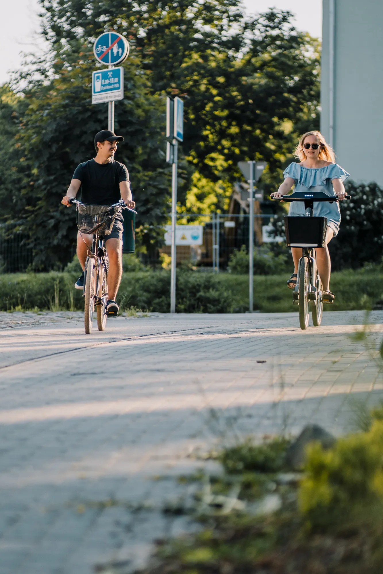 ** Kaks noort täiskasvanut sõidavad päikeselisel päeval Tartu linnas jalgratastel. Stseen näitab elavat linnaõhkkonda, tuues esile jalgrattateed ja rohelust, edendades jalgrattasõitu kui lõbusat ja keskkonnasõbralikku liiklemisviisi.