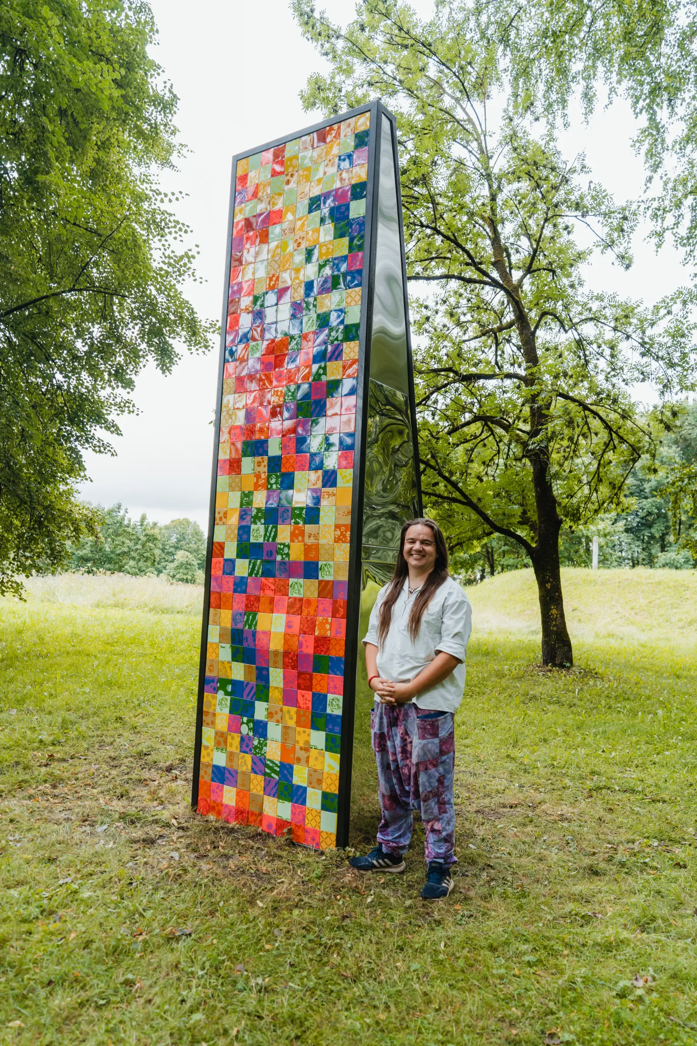 
Kõrge vertikaalne mosaiikpaneel värviliste ruutudega pargis. Naine seisab selle kõrval, taustal puud ja rohumaa.