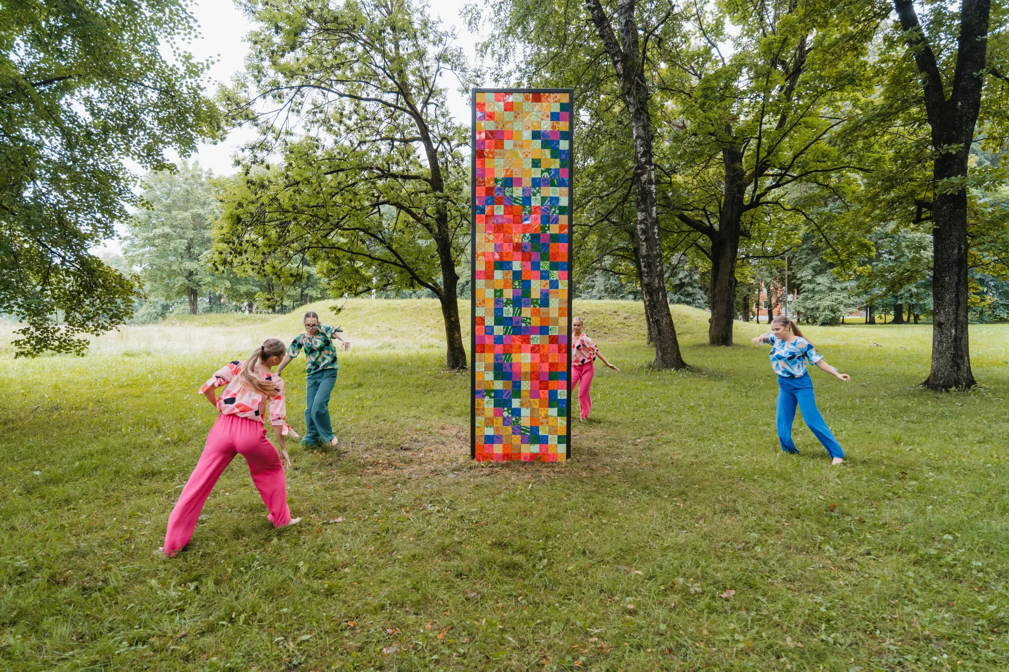  Päikesepaisteline Tartu park, kus seisab kõrge värviline mosaiikkolonn. Neli tantsijat erksates riietes tantsivad kunstiteose ümber rohelises pargis.