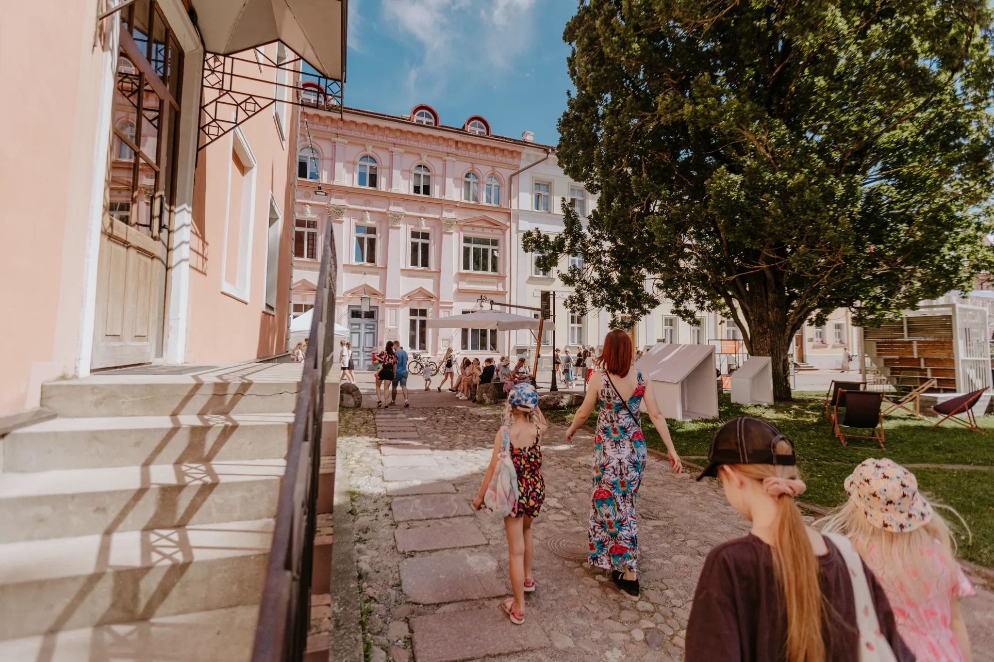  Naine ja lapsed kõnnivad Tartu vanalinna väljakul, kus on ajaloolised hooned, rohelus ja istumiskohad.