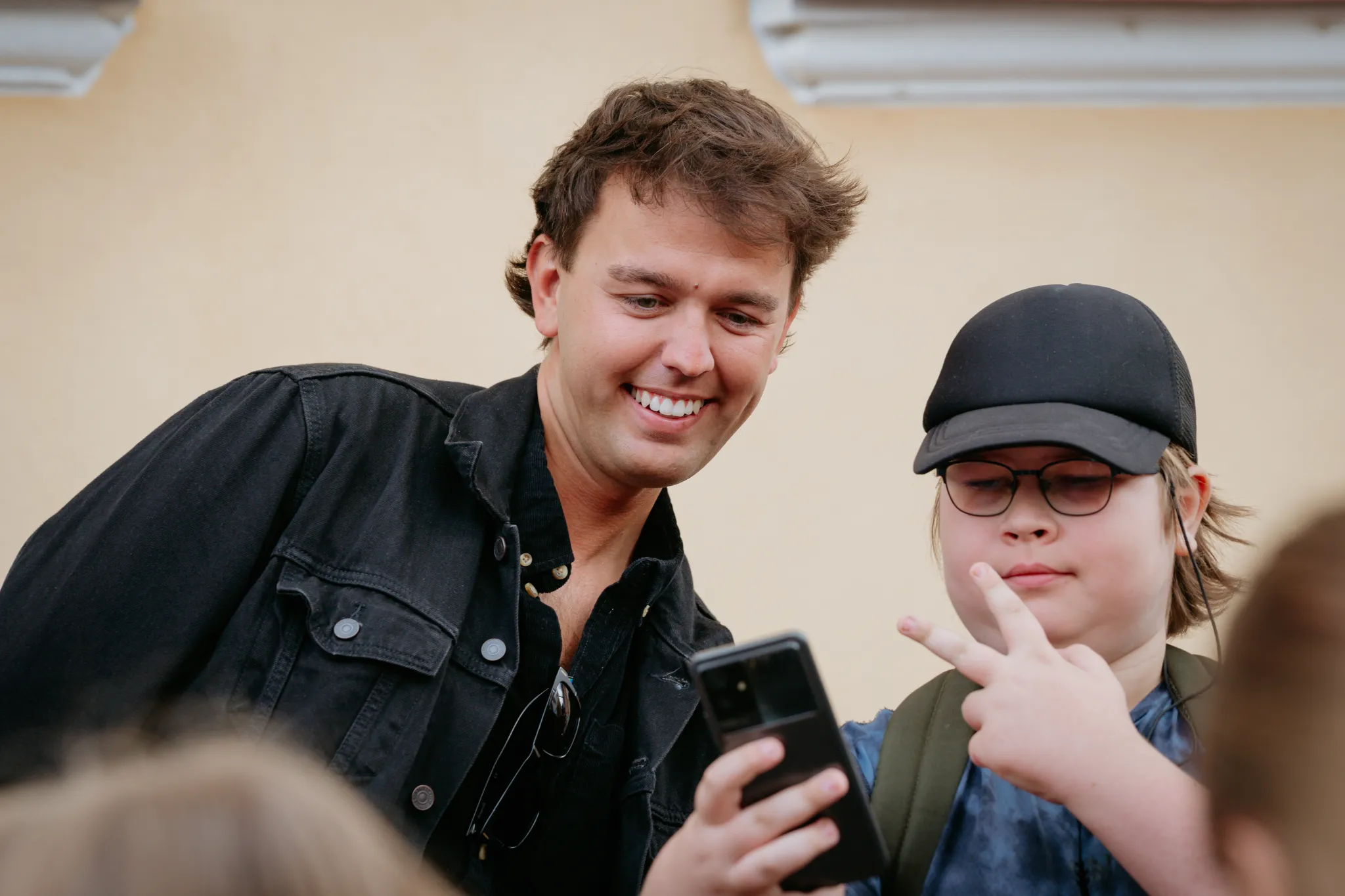  Naerusalus mees musta denim-jakiga teeb selfi noore fänniga mütsiga, vaatavad koos telefoni. Helge päeva tänavapilt.