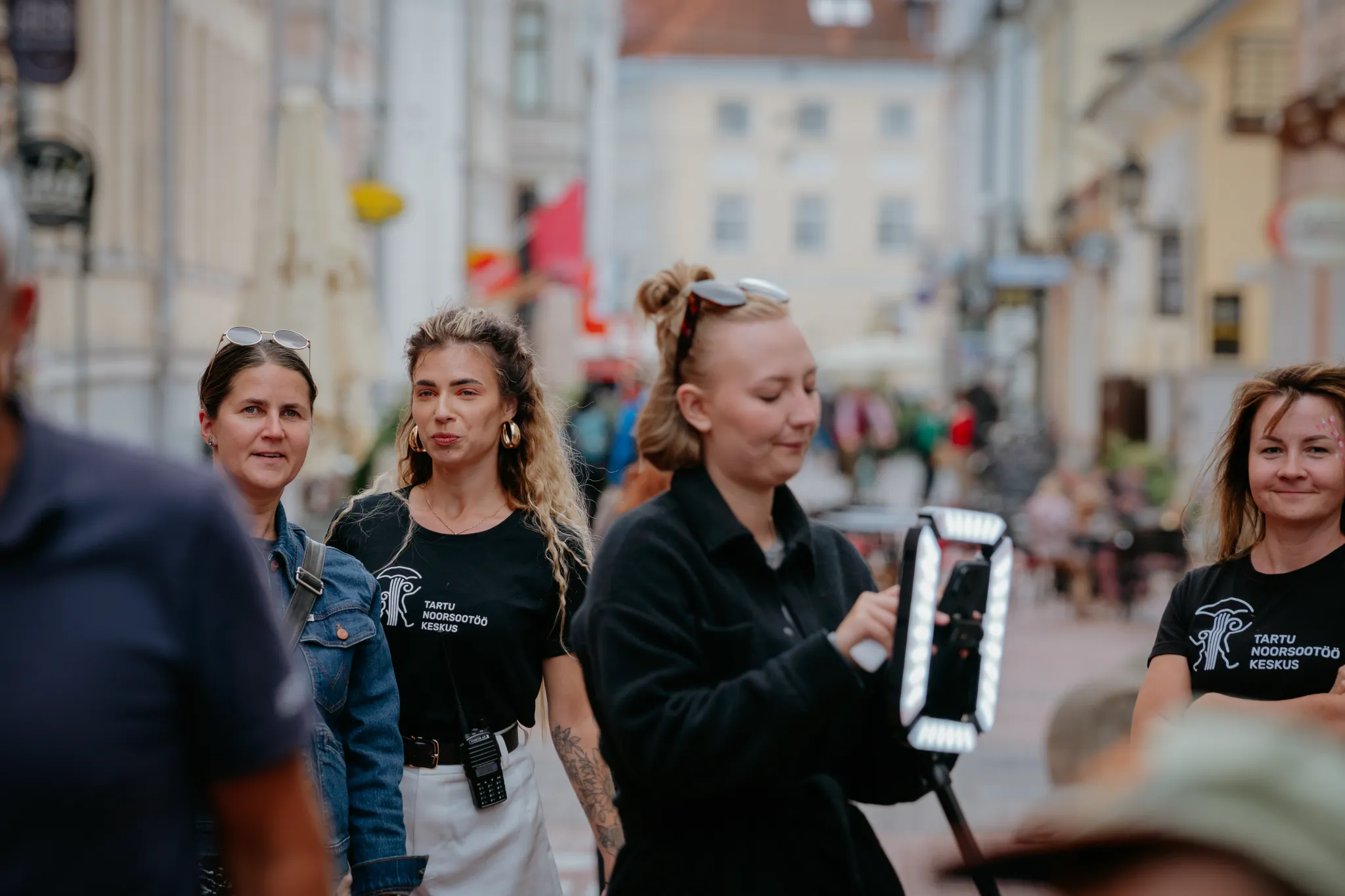  Tartu tänavavaade sõbralike giidide ja turistidega. Neli naist seisab munakivistil tänaval, üks kasutab valgustatud seadet giidiga kokemuseks. Soe, elav atmosüst Tartu ajaloolises keskväljapildis.