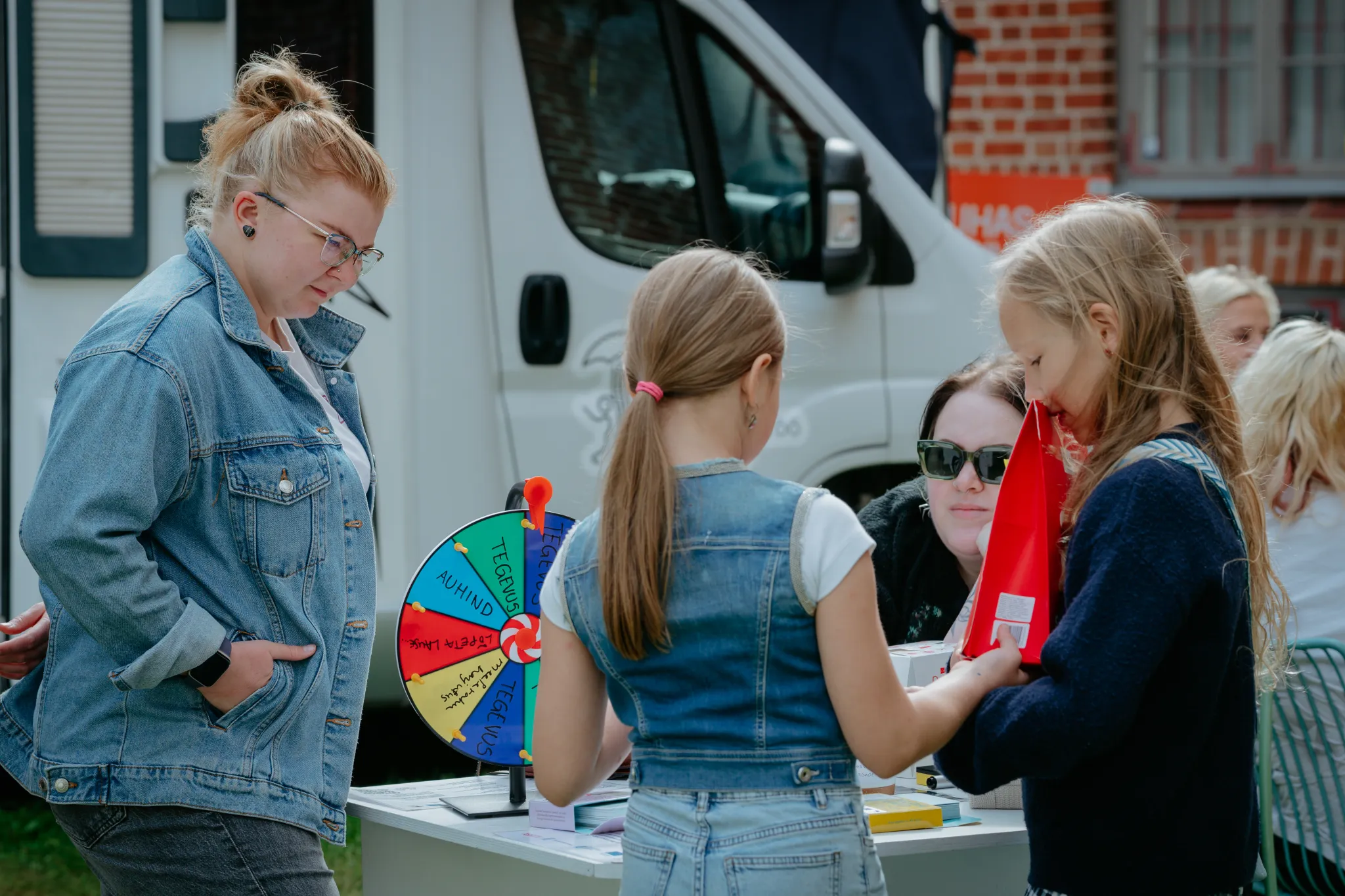  Inimesed välitelgi üritusel laua ümber, vasakul naine denimjakiga jälgimas; kaks last kannab denimriideid ja uurib punast eset. Taustal valgeanderja ja tellistest hoone ning van.