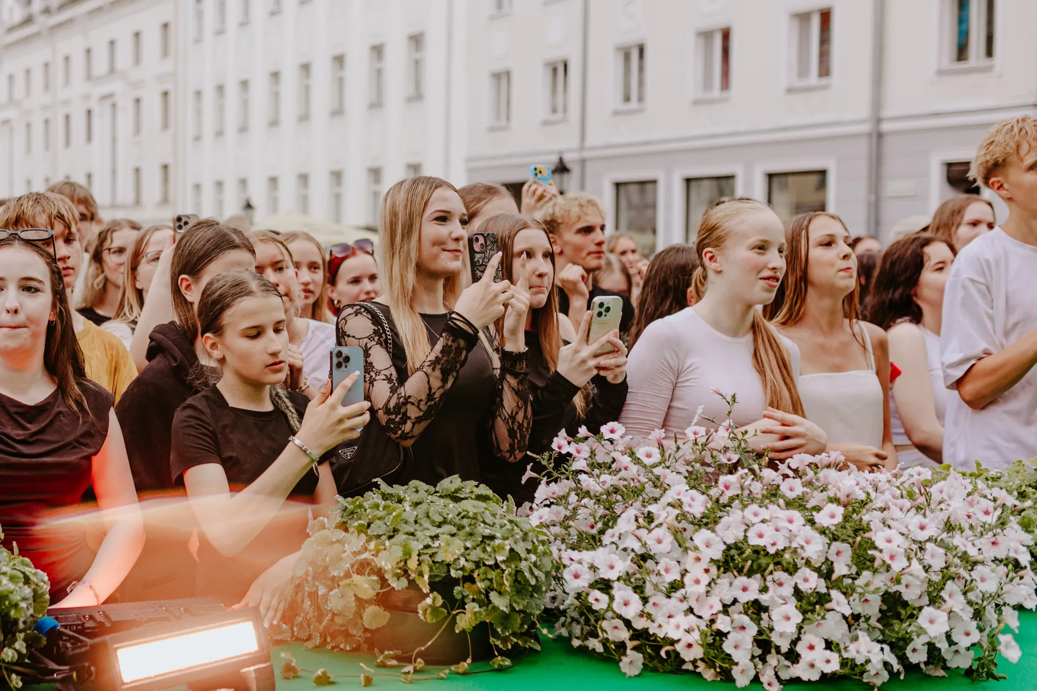  Põhjameed publik noortest inimestest järjekorras tänaval, paljud pildistavad telefonidega. Esiplaanil rohekates ja lillelistel dekoratsioonidel triiust on lilled ja auto/ekraan.