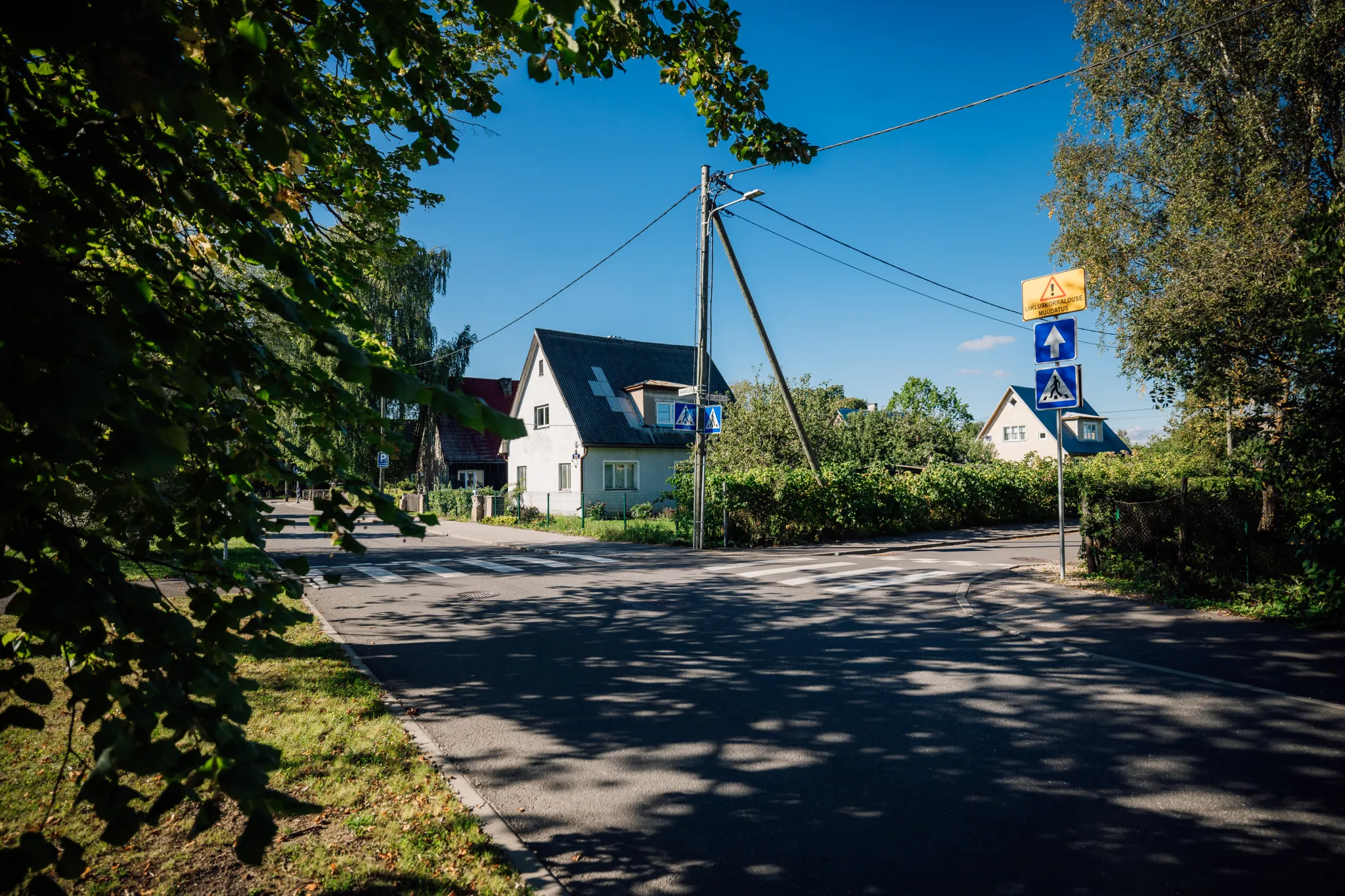  Päikesepaisteline elurajooni tänav. Vaikne ristmik ääristatud valgete kahekorruseliste majadega. Kalduv elektripost ja traadid. Puude varju, rohelised hekid ning hele sinine taevas loovad rahuliku, maalilise linnaosa meeleolu. Jalakäijate ülekäigumärgid 