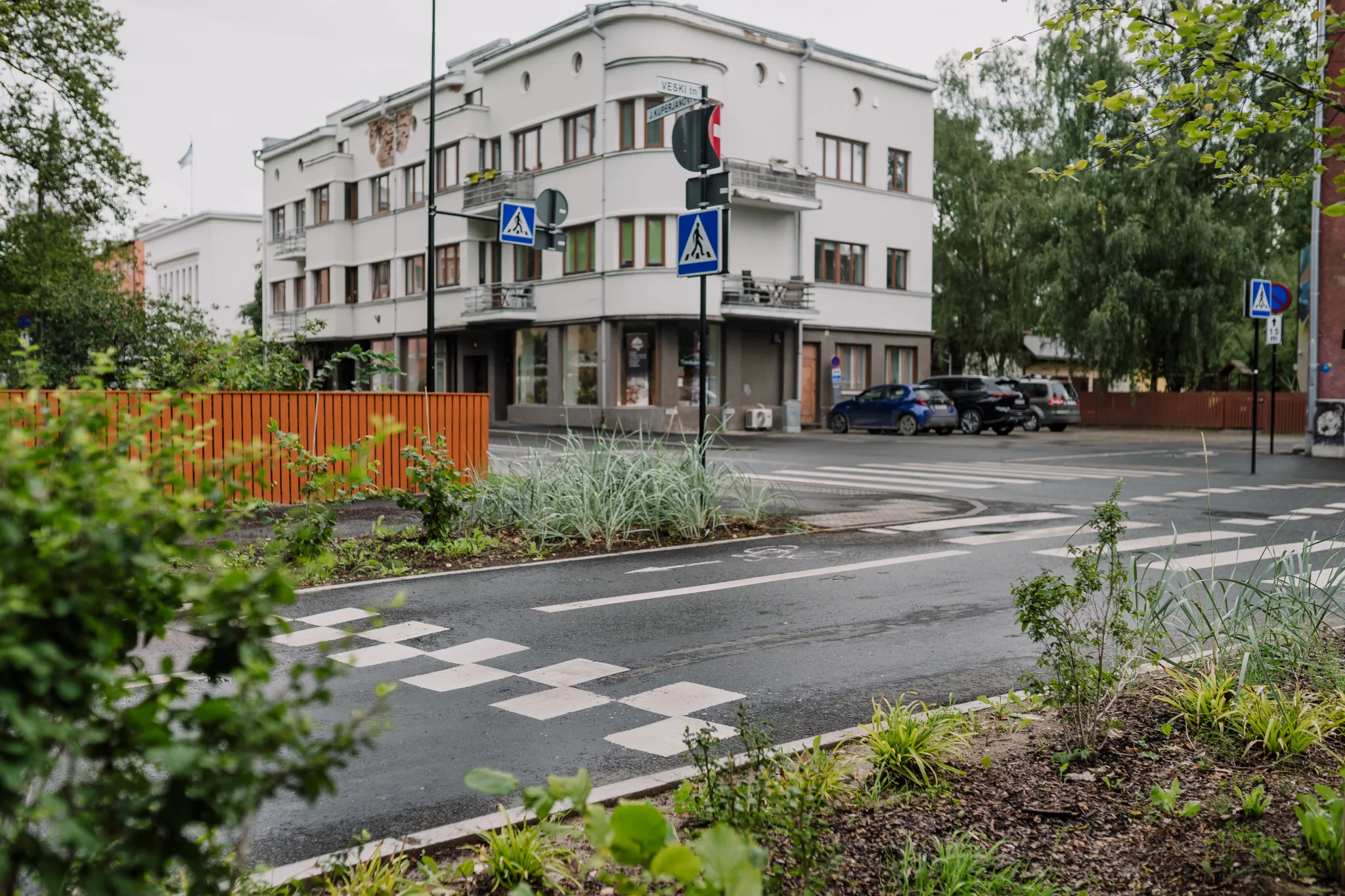 Vaade Veski-Kuperjanovi tänava ristmikule. Valge kolmekorruselise hoone ees on ülekäigurada ja liiklusmärgid. Tänava ääres kasvavad puud ja haljastus ning hoone kõrval on parkivad autod.