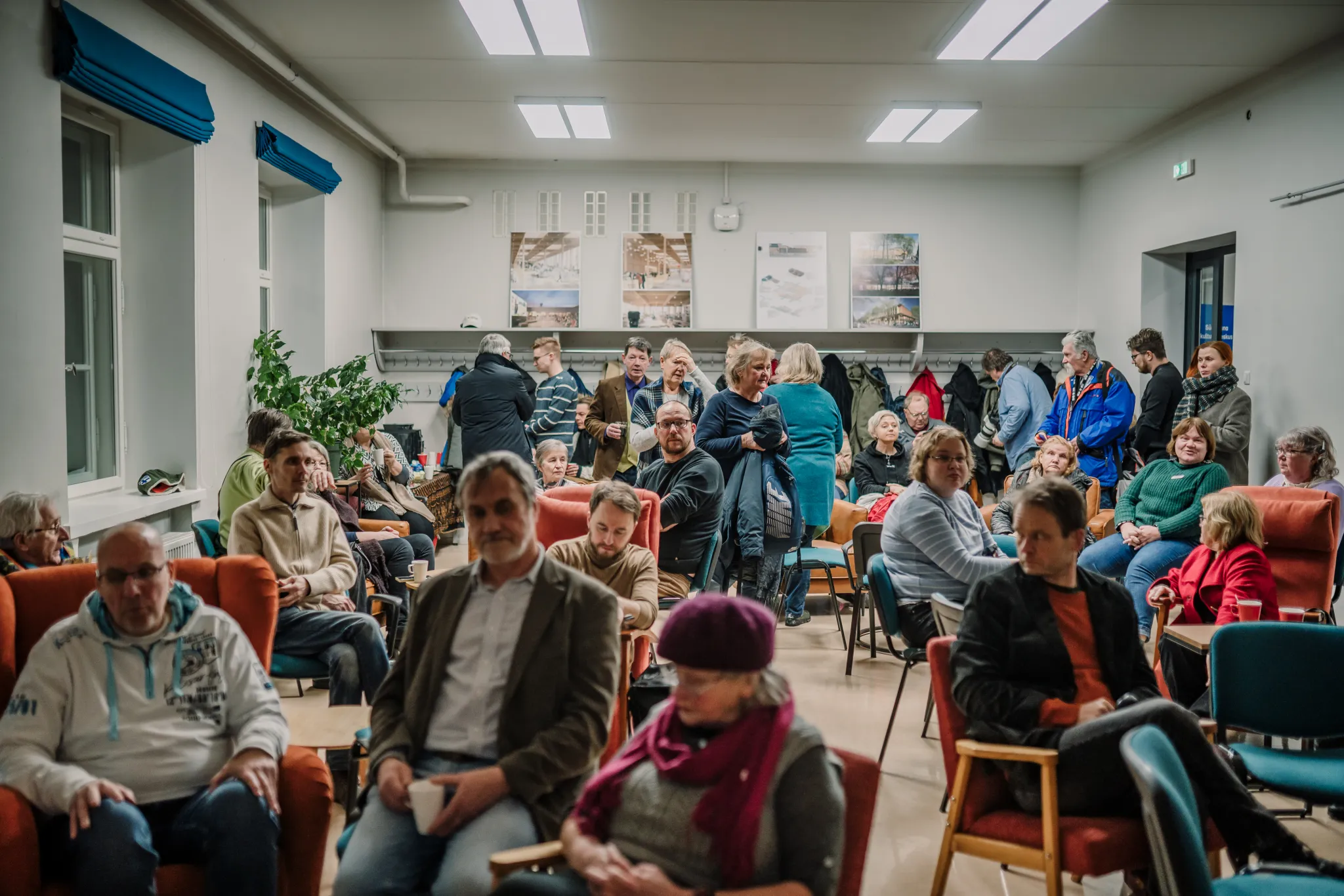 ** Rahvast täis kogukonna saal, kus istuvad osalejad, mõned vestlevad. Kohal on eri vanuses inimesi, luues elava atmosfääri. Seintel on nähtavad plakatid, mis viitab, et toimus üritus või koosolek.