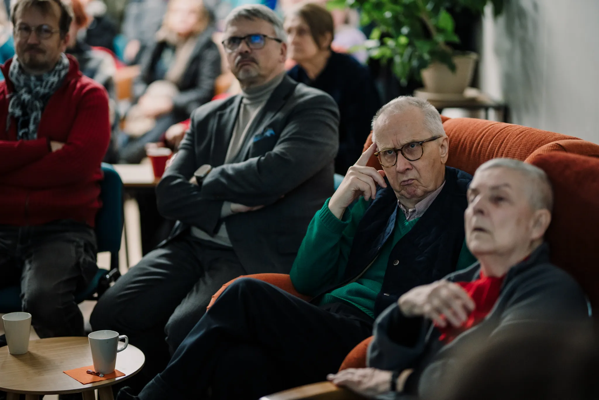 ** Rahvast täis kogukonna saal, kus istuvad osalejad, mõned vestlevad. Kohal on eri vanuses inimesi, luues elava atmosfääri. Seintel on nähtavad plakatid, mis viitab, et toimus üritus või koosolek.