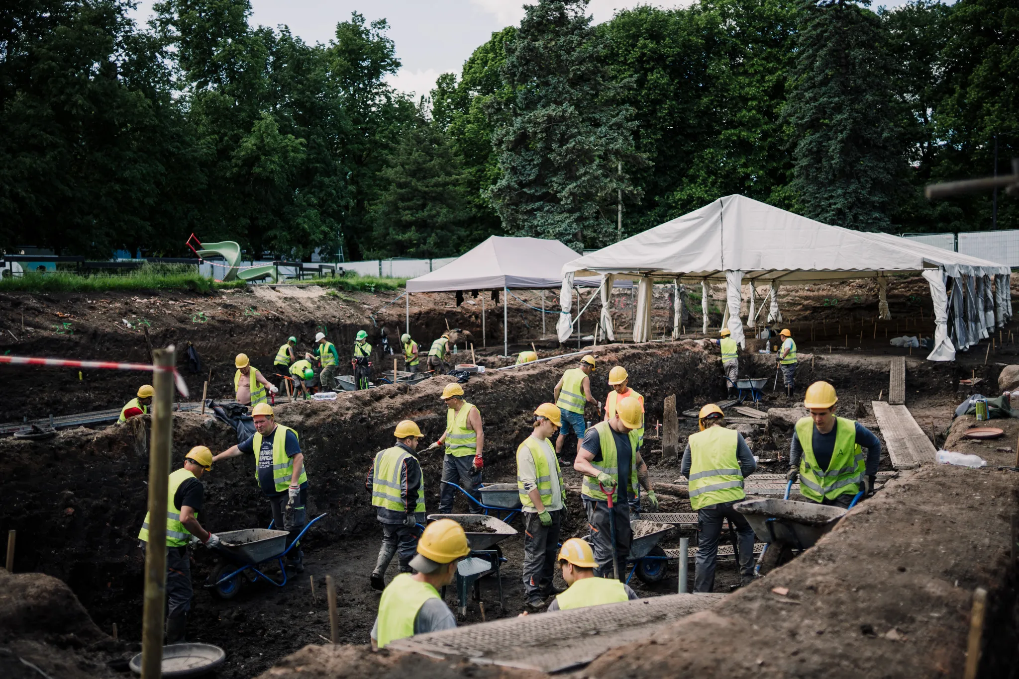  Ehitusplatsil palju töötajaid kollaste kiividega ja heledate vestidega, kaevatakse kaevikuid valgete telkide all; taustal puud ja roheline park.