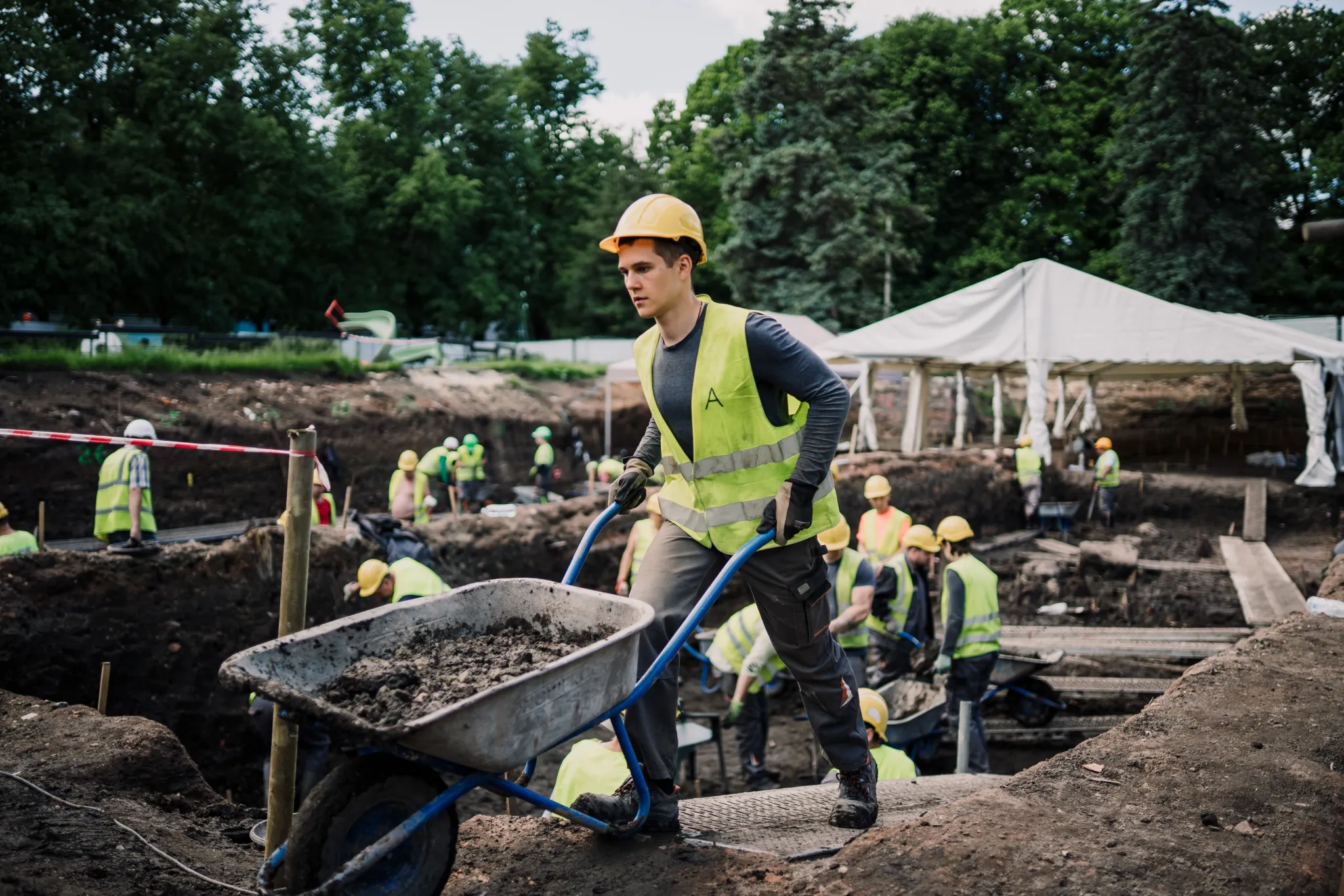  Pargi ehitusplats: töölised kollaste helkurvestide ja kiivritega liigutavad mulda käruga; taustal telgid ja ehitustöö meeskond.