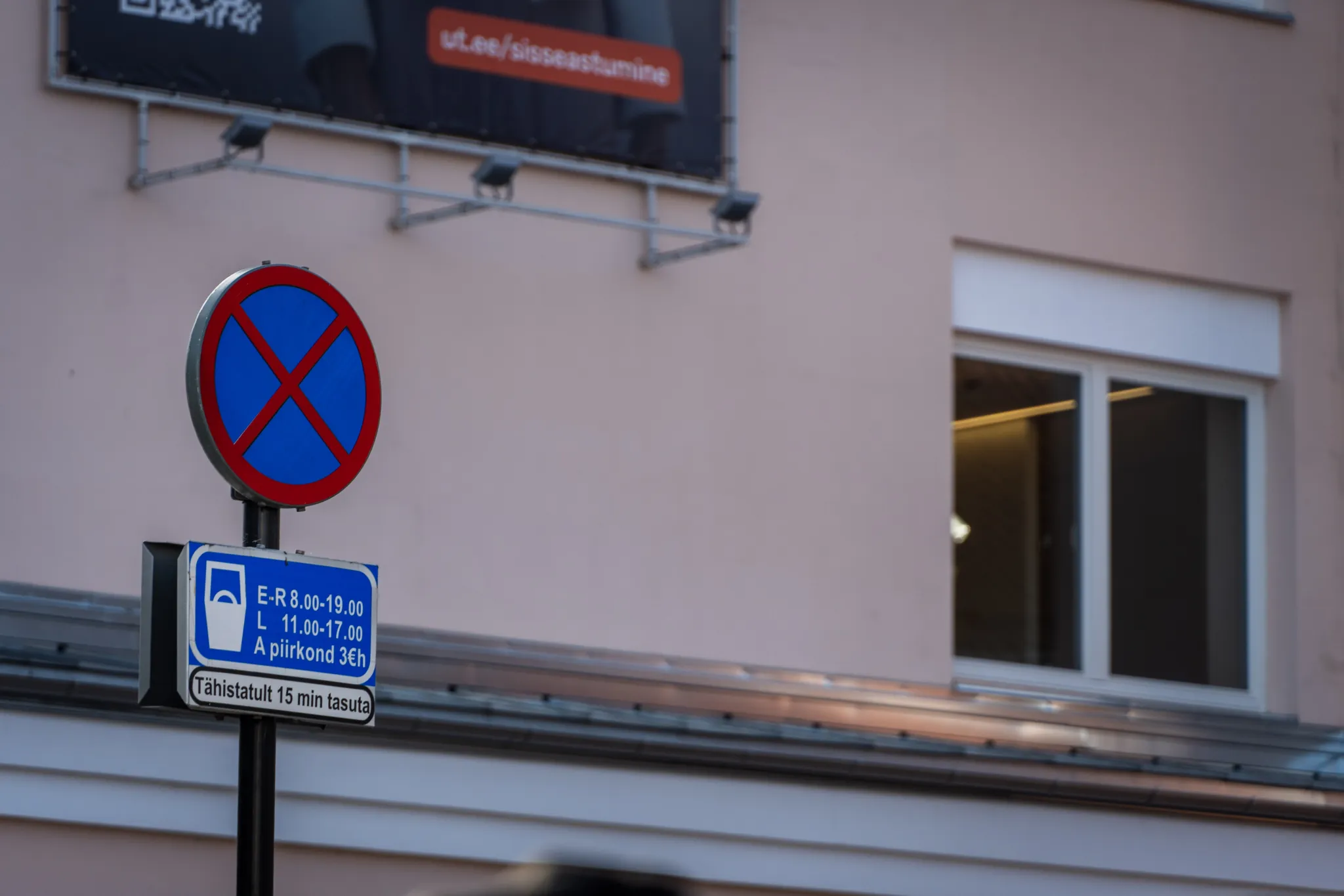  Linna tänava stseen: sinine peatust keelav silt postil ja all ajalimiidiga parkimis tahvel. Taustal roosa hoone ja valgustatud aken.