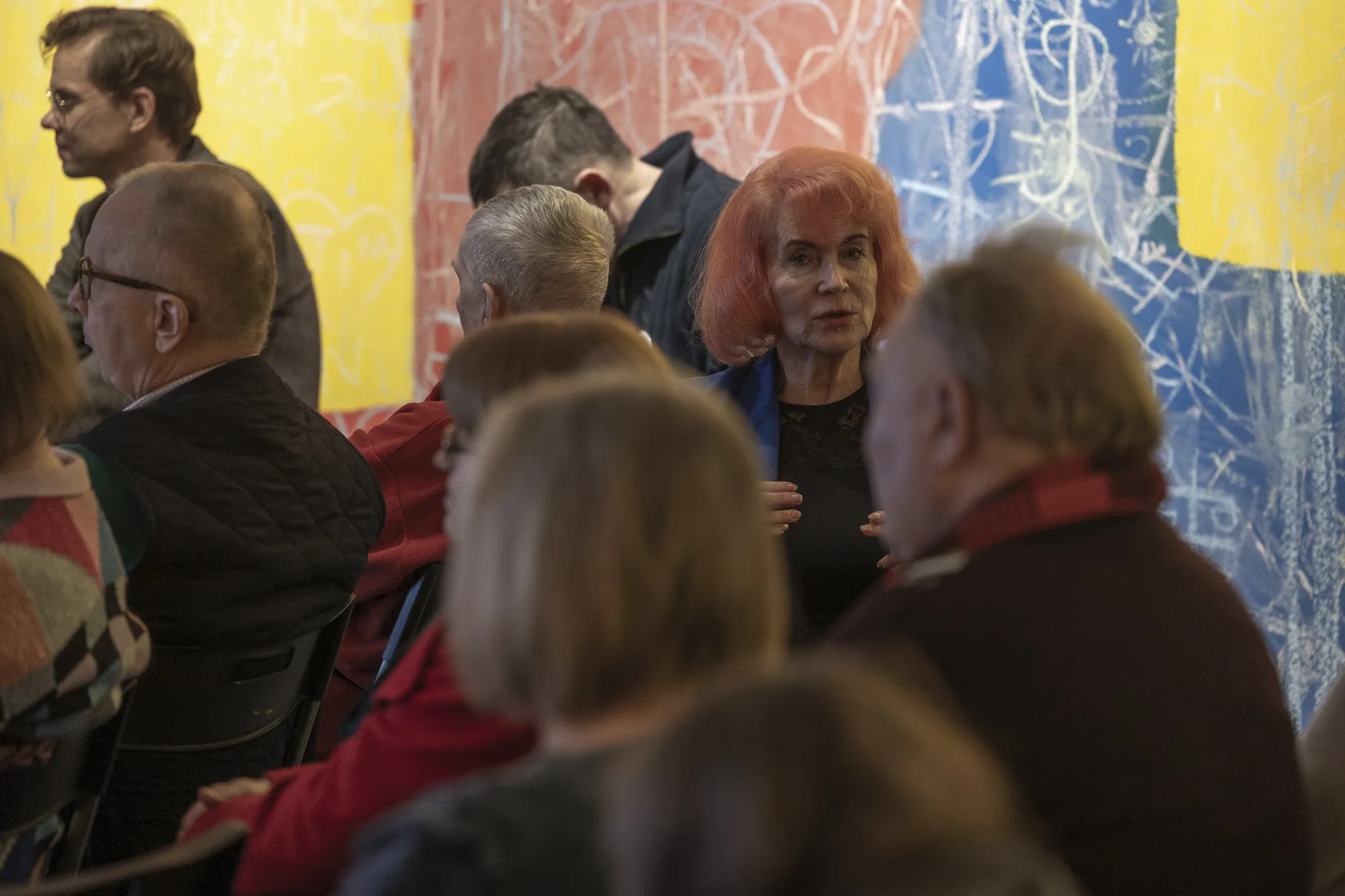 ** Tartu üritusel koguneb värvikas publik. Esiplaanil suhtleb erkhaiguse naine külalistega. Taustal on värvikad abstraktsed kunstiteosed, mis lisavad üritusele elavat atmosfääri.
