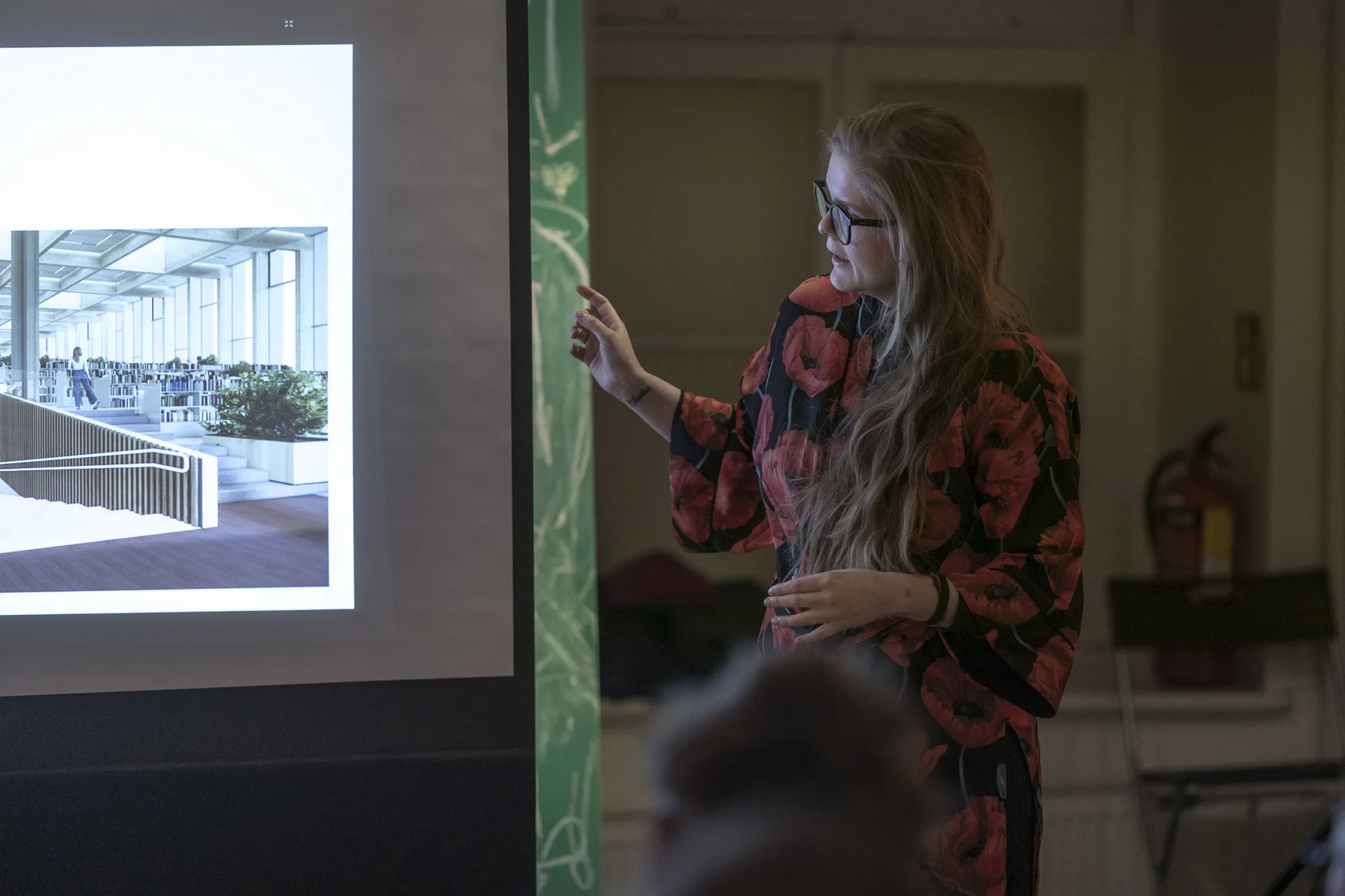 ** Erineva taustaga publikuliige silmapaistva soenguga kuulab tähelepanelikult sinise kriidiseina ees, ümbritsetud teistest vestlevatest osalejatest. Stseen peegeldab Tartu kogukonnaürituse elavat atmosfääri.