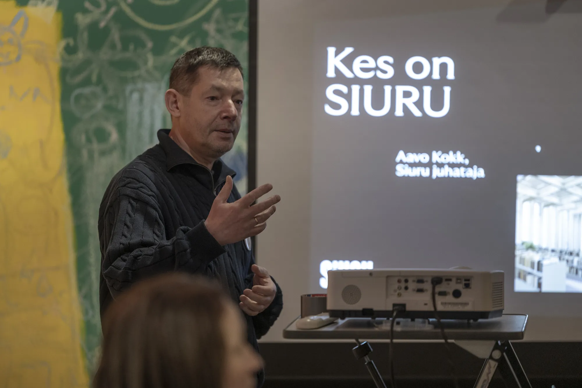 ** Mees seisab esitluse ekraani ees, arutades "Kes on SIURU?" Ta jutustab ja kasutab žeste, kaasates publiku. Taustal on värviline seinamaaling. Üritus rõhutab suhtlemist ja teabe jagamist Tartus.