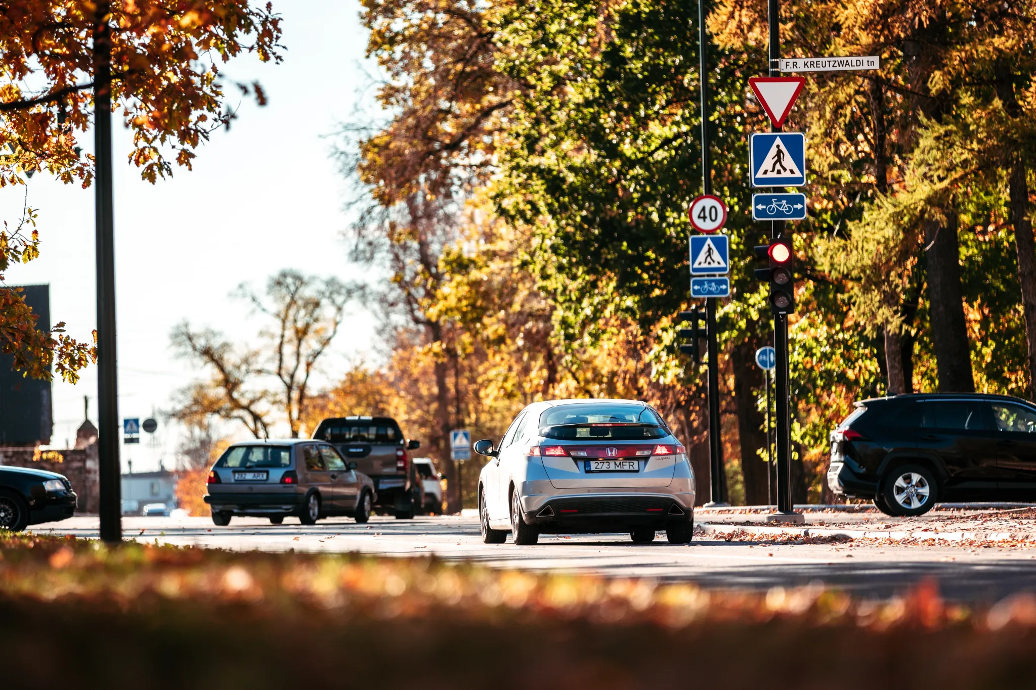 Autod sõidavad sügisesel Tartu tänaval kuldsete puude ja liiklusmärkide all.