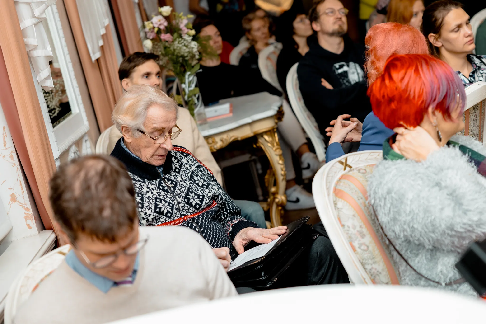 ** Mugav koosolek Tartus, kus on kaasahaarav publik. Erinevad inimesed, sealhulgas eakam mees mustrilises kampsunis, istuvad ja kuulavad tähelepanelikult. Õhkkond on soe ja kutsub, ideaalne kogukonnaürituste ja kultuuriliste arutelude jaoks.