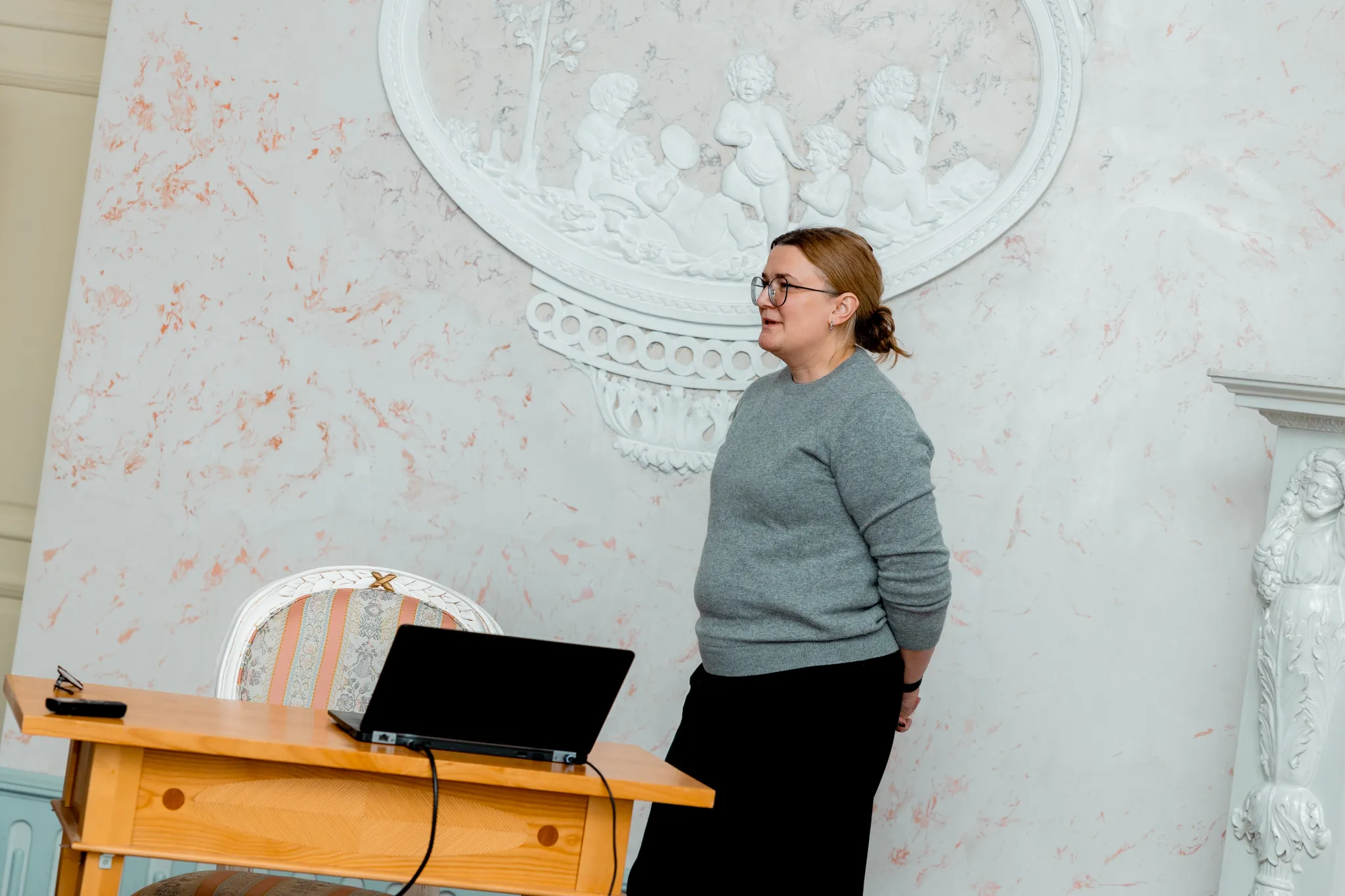 ** Noormees mustas ülikonnas näitab esitlusel žeste, seistes elegantses toas. Puust laual on avatud sülearvuti. Taustal on laste kujutisega dekoratiivne seinakunst. Keskkond edastab professionaalset atmosfääri.