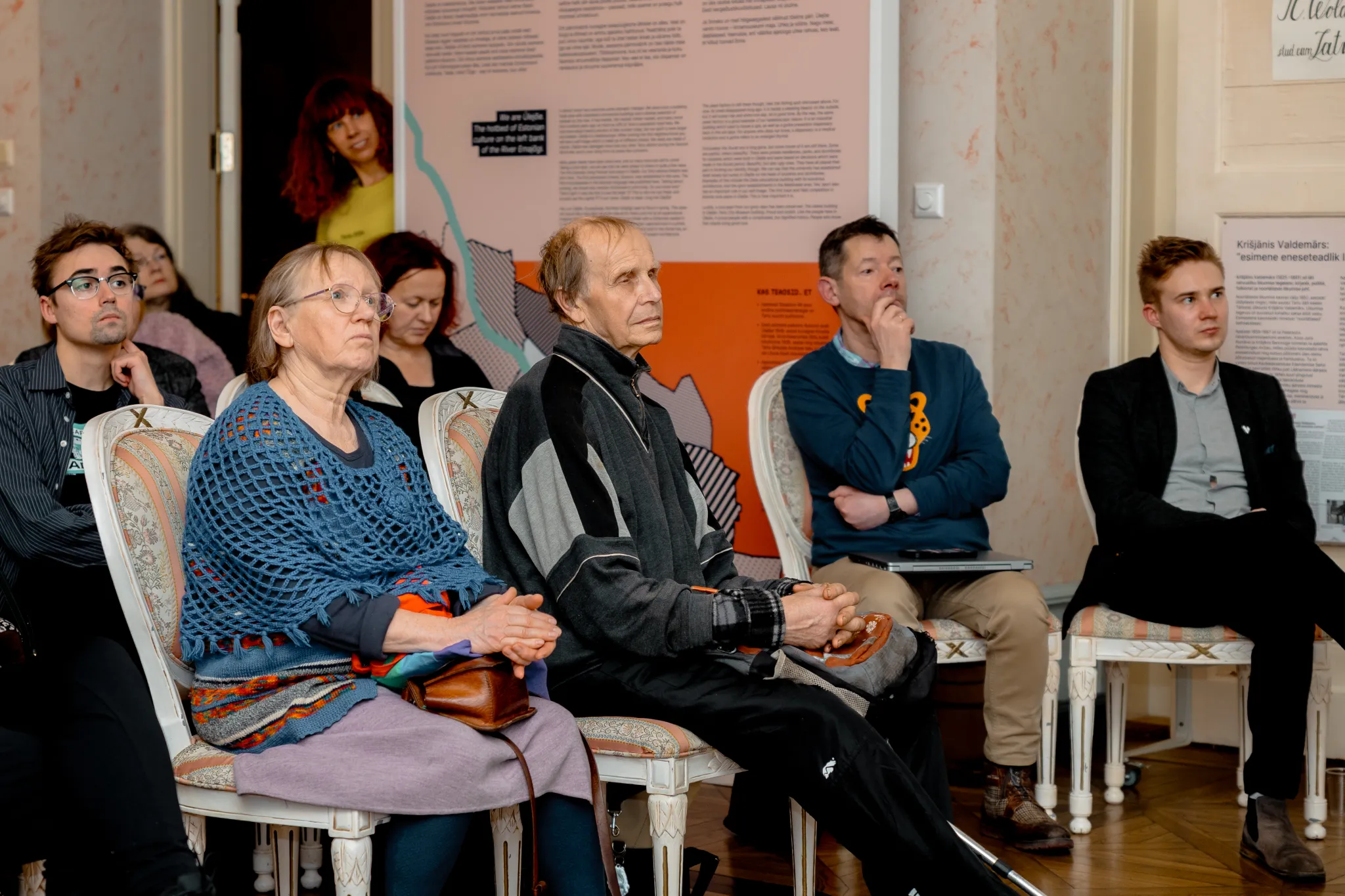 ** Mitmekesine ja huvitatud publik istub tähelepanelikult valgusküllases ruumis. Nad näitavad erinevaid reaktsioone, mis viitavad huvi tundmisele Tartu esitlusest või arutelust. Ruumi kaunistavad kunstilised seinalehted, mis lisavad atmosfäärile.