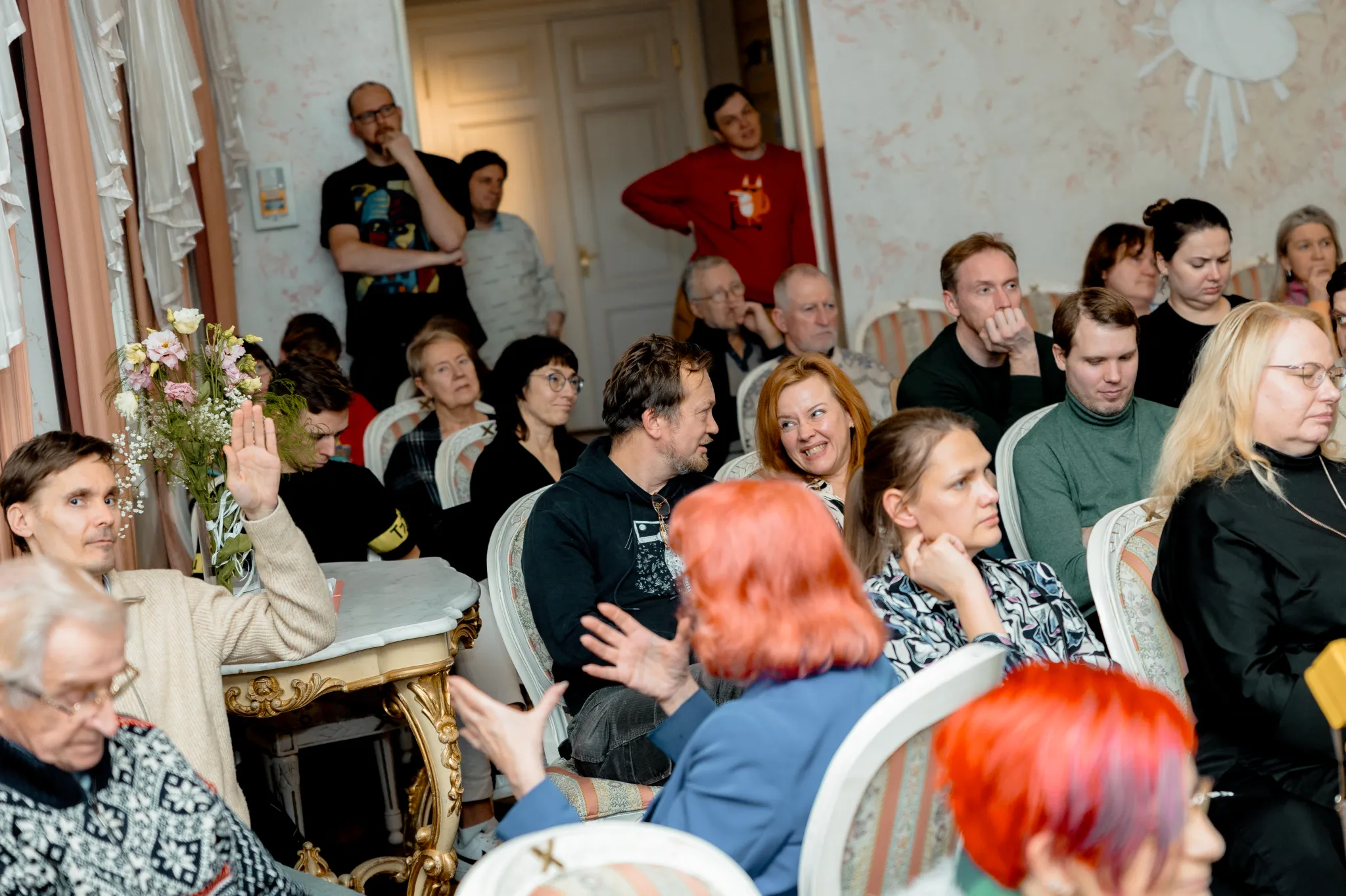 ** Mitmekesine osalejate grupp istub hubases toas, osaledes elavas arutelus. Mõned tõstavad käsi, et kaasa rääkida, teised kuulavad keskendunult. Ruumi iseloomustavad elegantsed toolid ja lillede kaunistused, luues külalislahke õhkkonna kogukonna suhtlemi