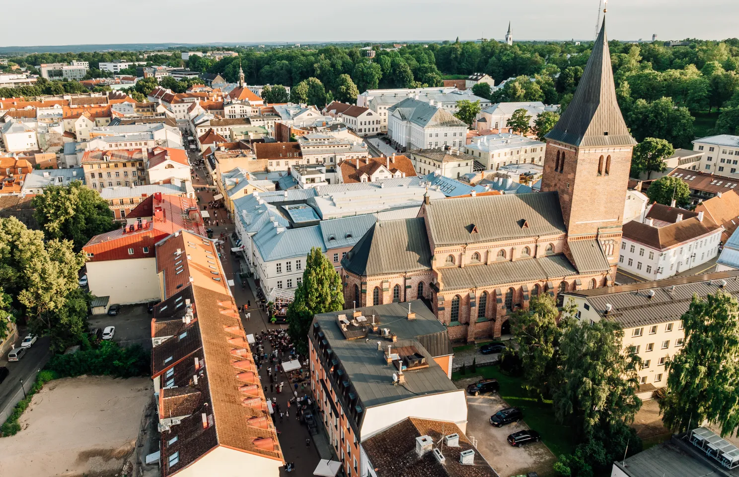  Ilmvaade ajaloolise Euroopa linna pealt: punaste katustega majad, suur paekirikutest kirik ja roheline taust. Tänavad täis inimesi ning värvikirevad hooned.