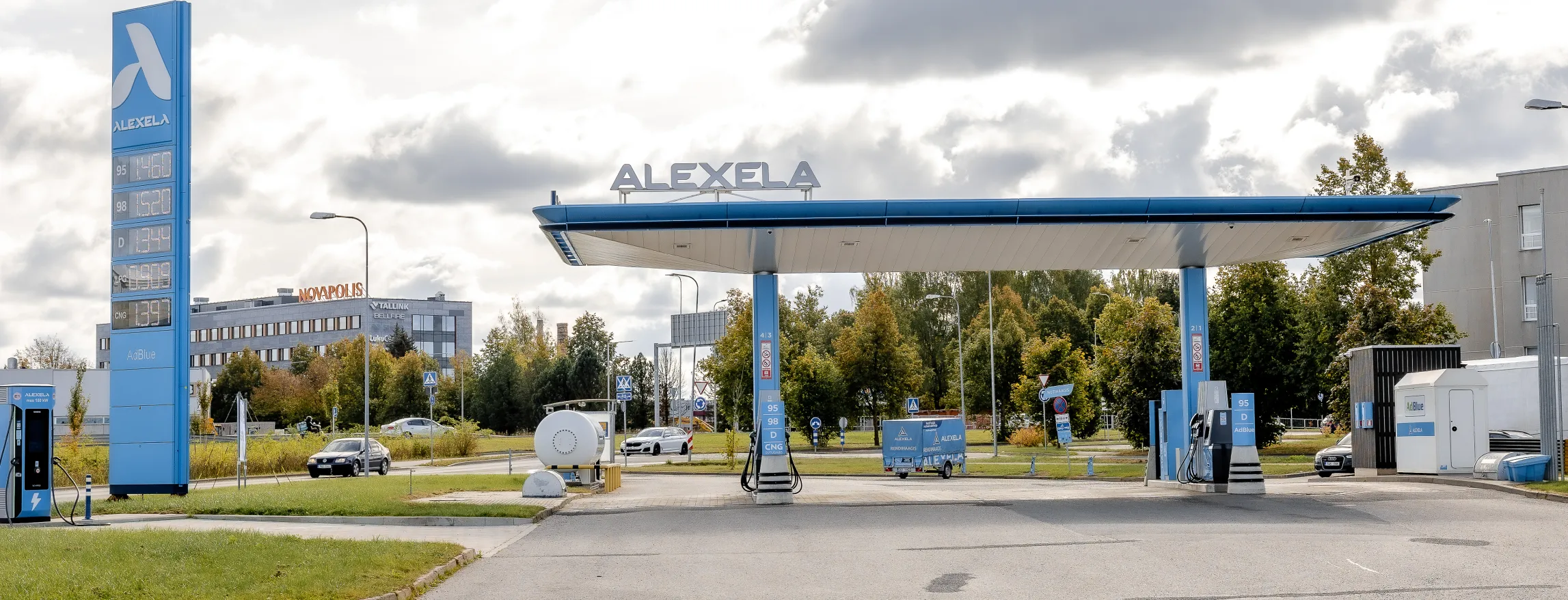  Alexela tankla sinise katuse ja märgistusega, kütusepumbad ees, autod teel, taustal puud ja pilved.