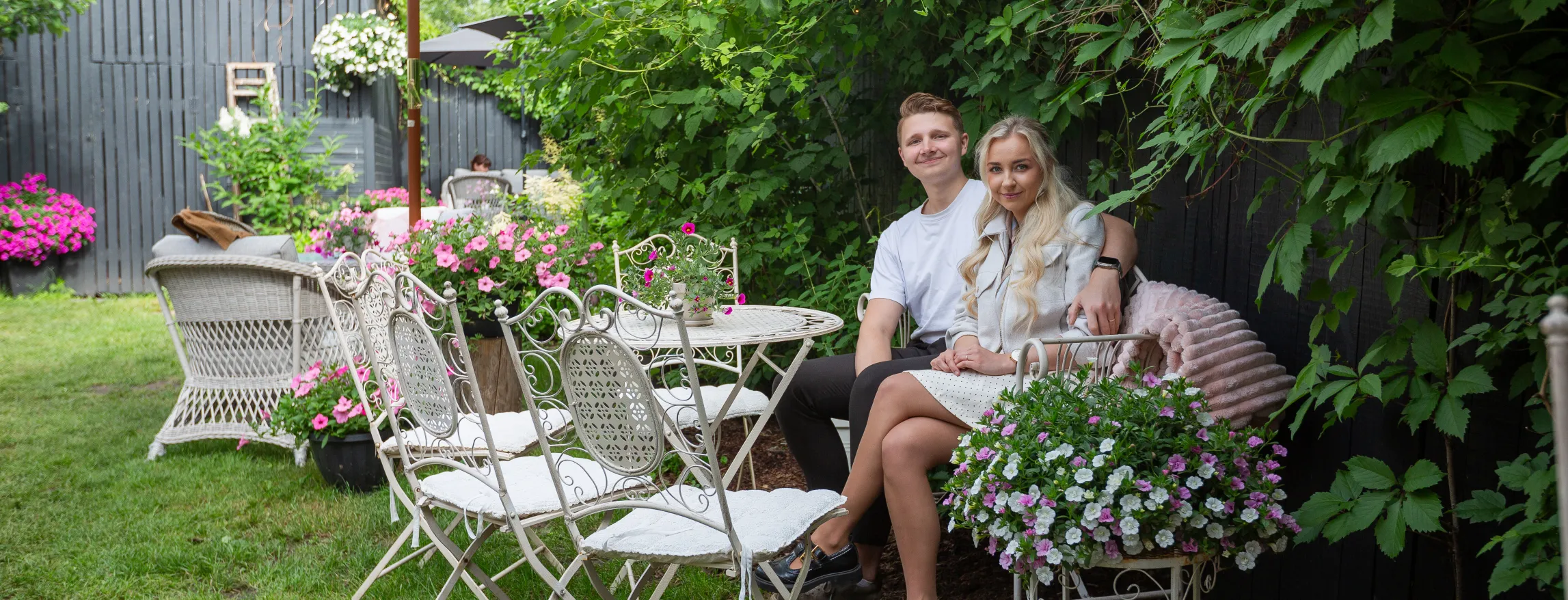  Paar istub valgete raudaiatoolide peal suurvalge päikesevarju all, lopsakas Eesti aed taustal. Pottlilled ja rohelised ronitaimed loovad hubase välimuse.