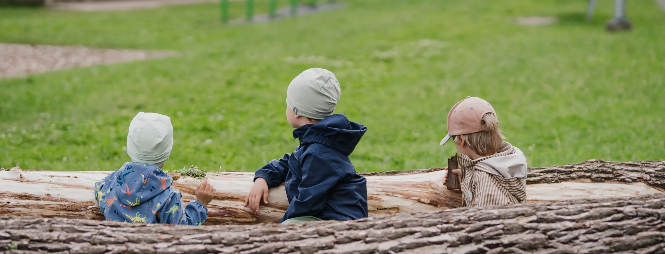  Kolm last istuvad suurel puupalgil pargi rohelisel murul. Nad on jakides ja mütsikesed ning vaatavad eemale. Taustal on mänguväljak ja rohelus.