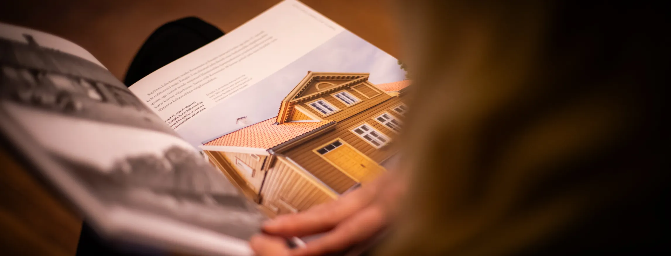  Lähedalt näha inimest, kes loeb reisibrošüüri; käed keeravad lehte ja sooja foto puidust majast katusega rõhutab arhitektuuri ja kohaliku võlu.