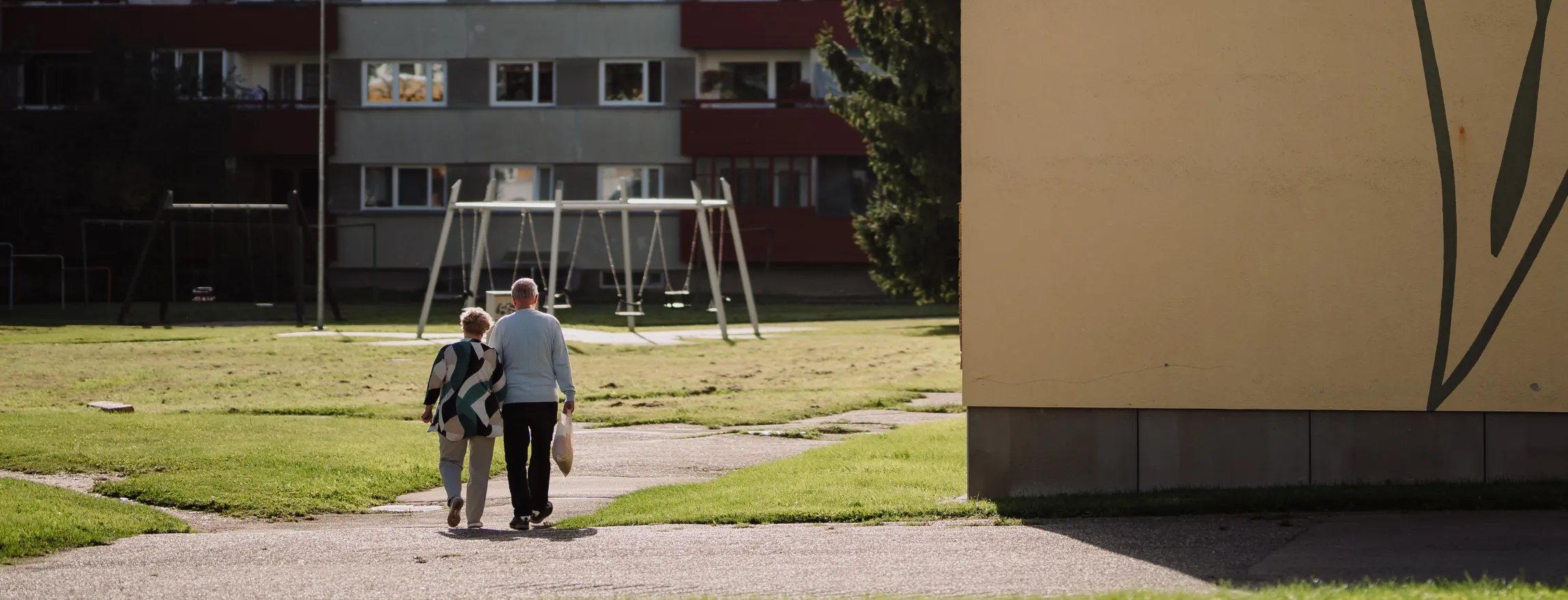 Linnaparklik vaade Tartust: vanem paar kõnnib käsikäes mööda teerada, taustal korterelamud ja mänguväljakukorv, päikeseloojangul soe valgus.