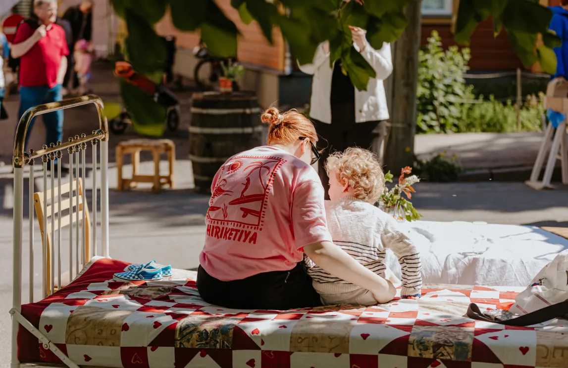  Väljakul päikeselises pargis istub roosas särgis inimene voodil, millel on lapitekist patchwork. Kõrval on väike laps; taustal vabatahtlikud ja pealtvaatajad.