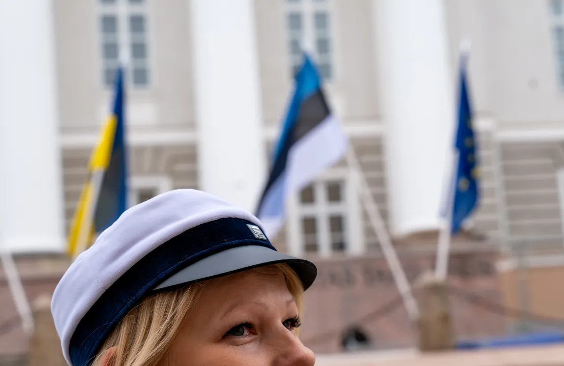  Naine valge kapteni mütsiga vaatab üles. Taustal on valged sambad ja lipud, mis tähistavad avalikku hoonet.