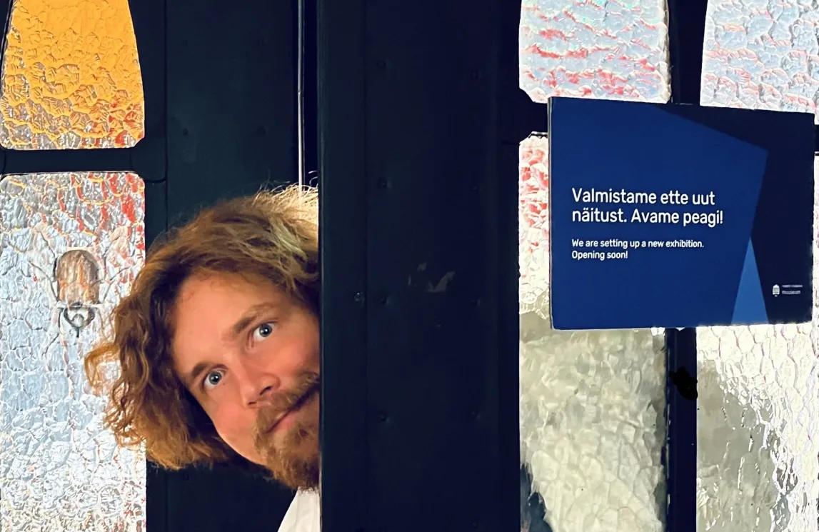 Inimene piilub musta seina tagant, käes aju makett. Taustal on reljeefsed pinnad ja silt „Valmistame ette uut näitust. Avame peagi!“.